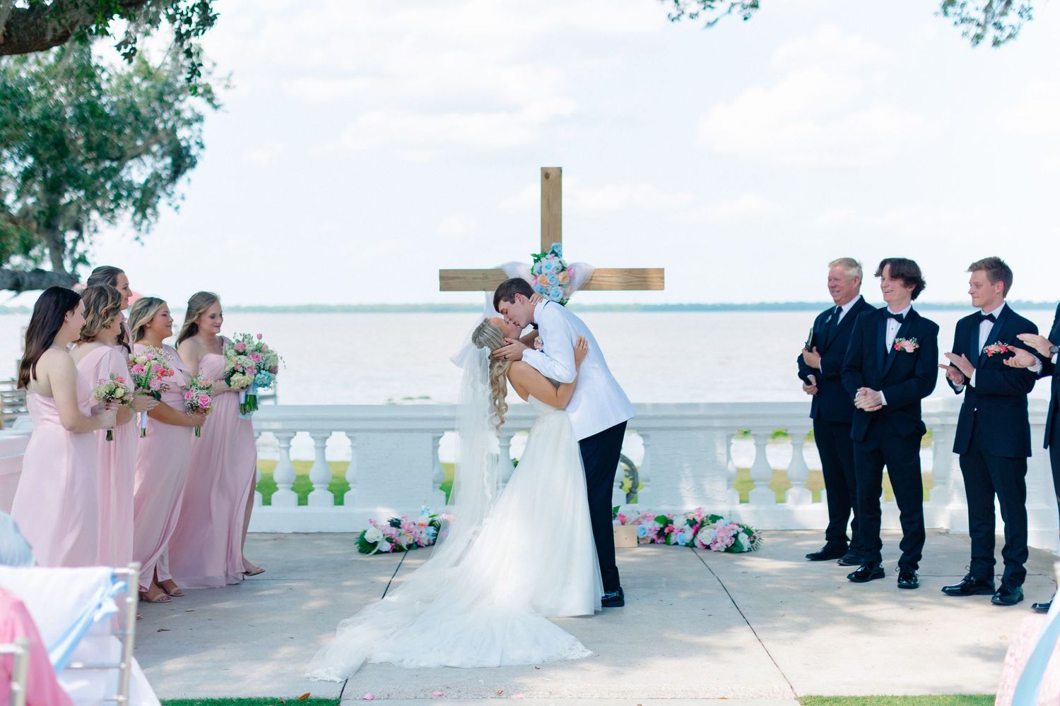 Bride and groom share first kiss at outdoor waterfront wedding ceremony with bridesmaids in pink and groomsmen in black suits, Florida
