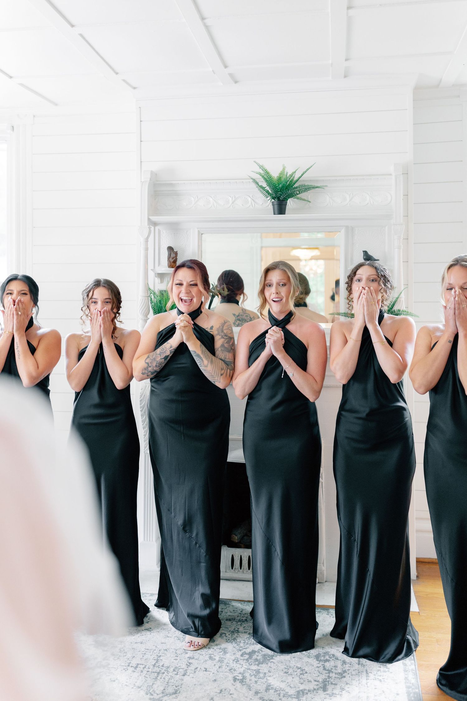 Bridesmaids in black halter gowns react with joyful surprise seeing the bride, hands raised to mouths in a white venue.