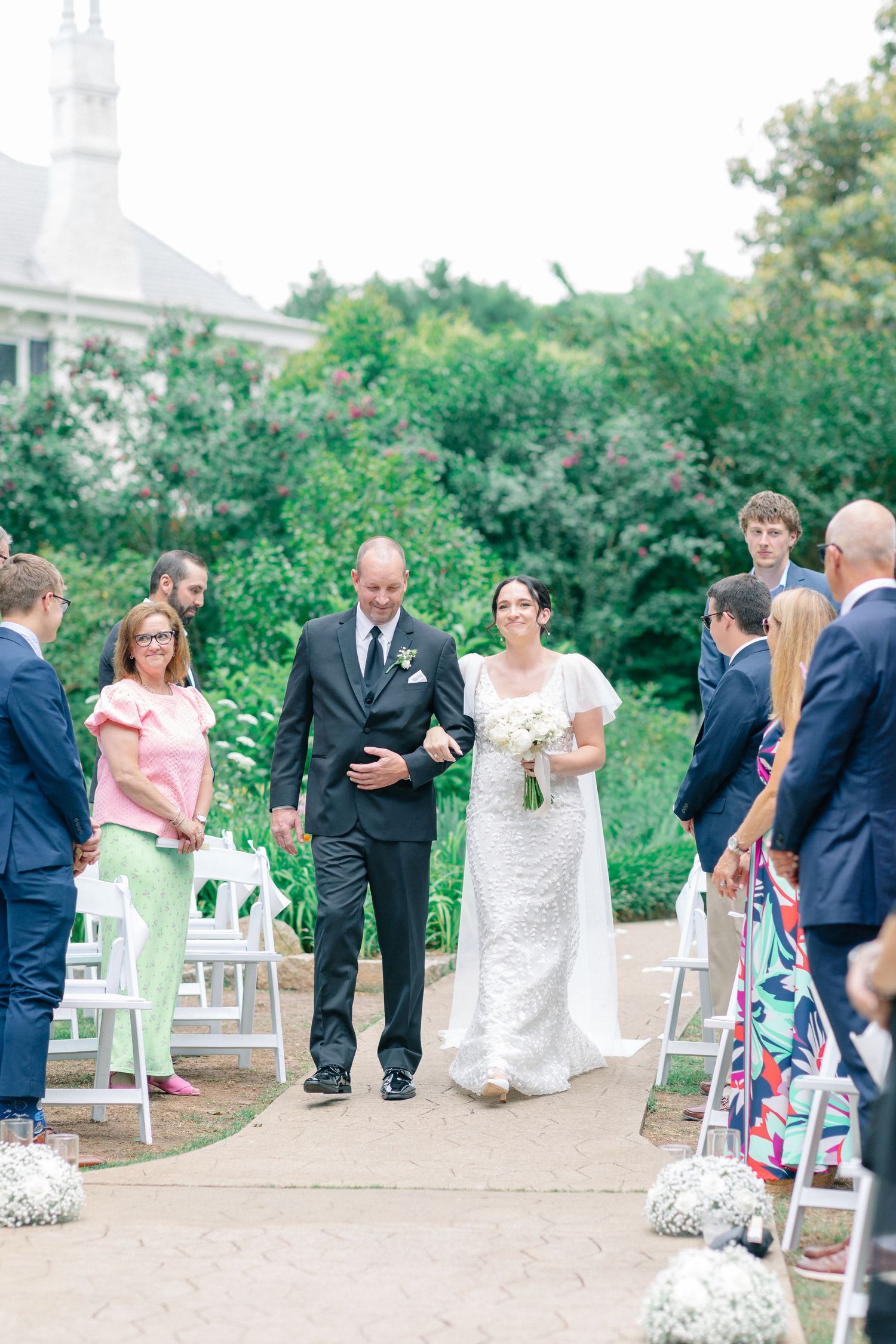 Bride walks down outdoor aisle with escort, guests standing, lush greenery backdrop, elegant white lace gown, bouquet.