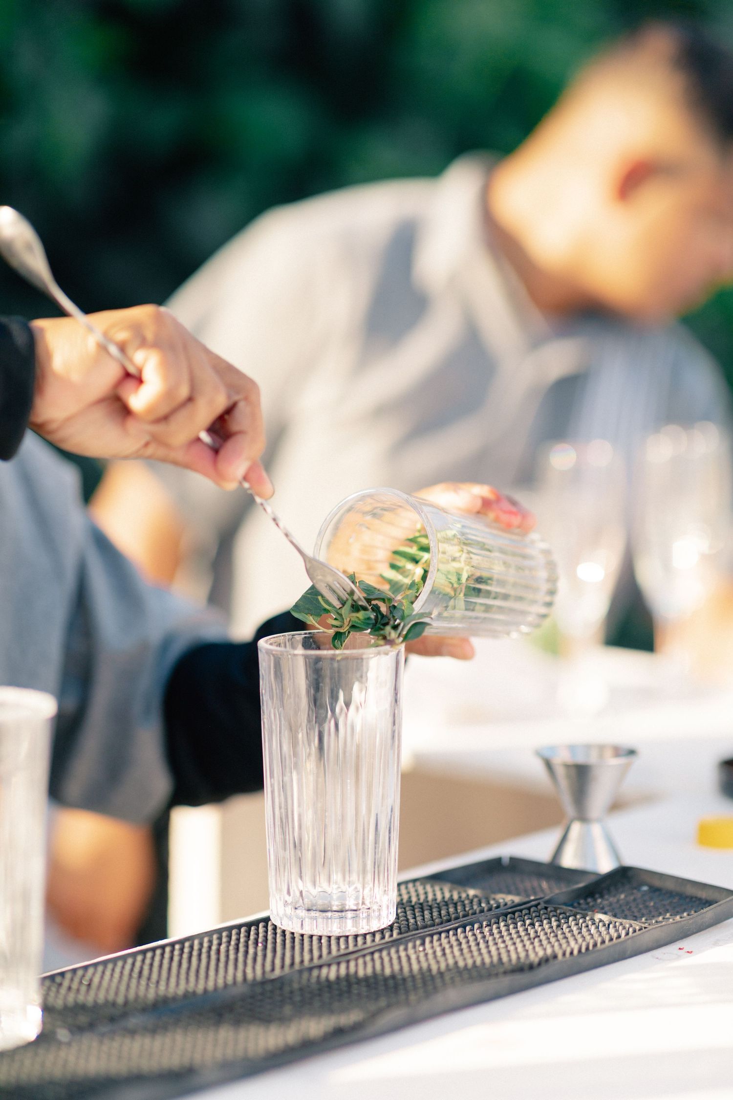 Bartender carefully transferring fresh herbs into a crystal highball glass on a bar mat at an outdoor event.