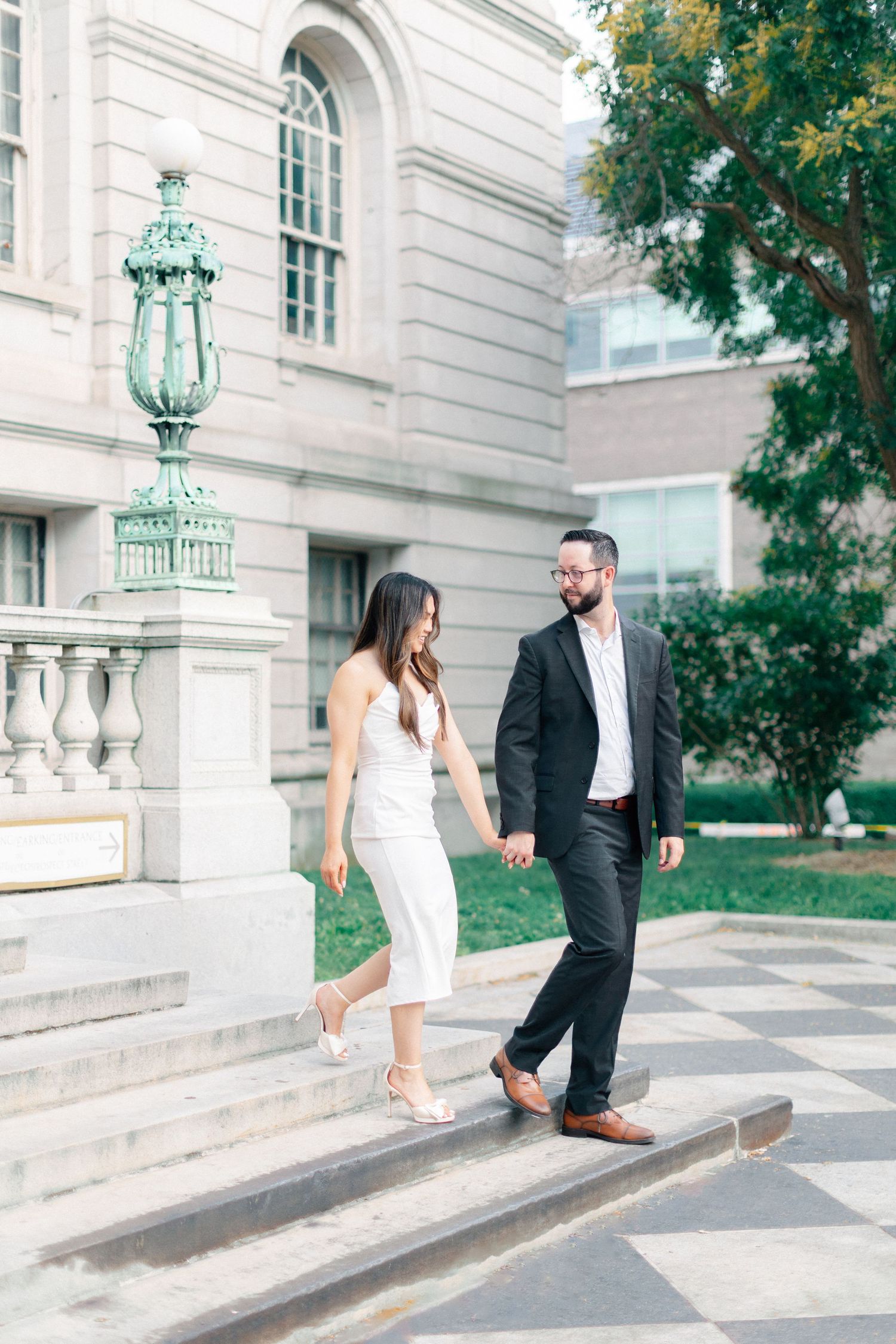 Elegant couple holds hands walking down stone steps outside a classic beaux-arts building with green lamp post.