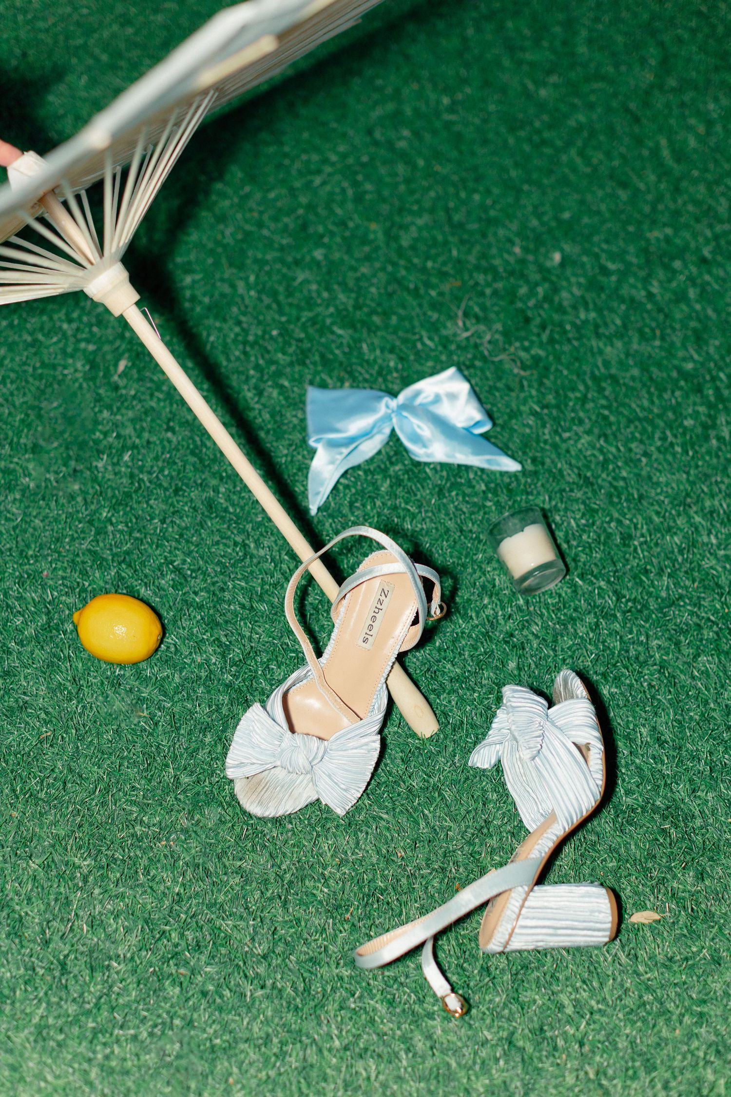 White bow heeled sandals on green turf with a blue satin bow, lemon, candle, and decorative parasol, wedding at Bella Cosa