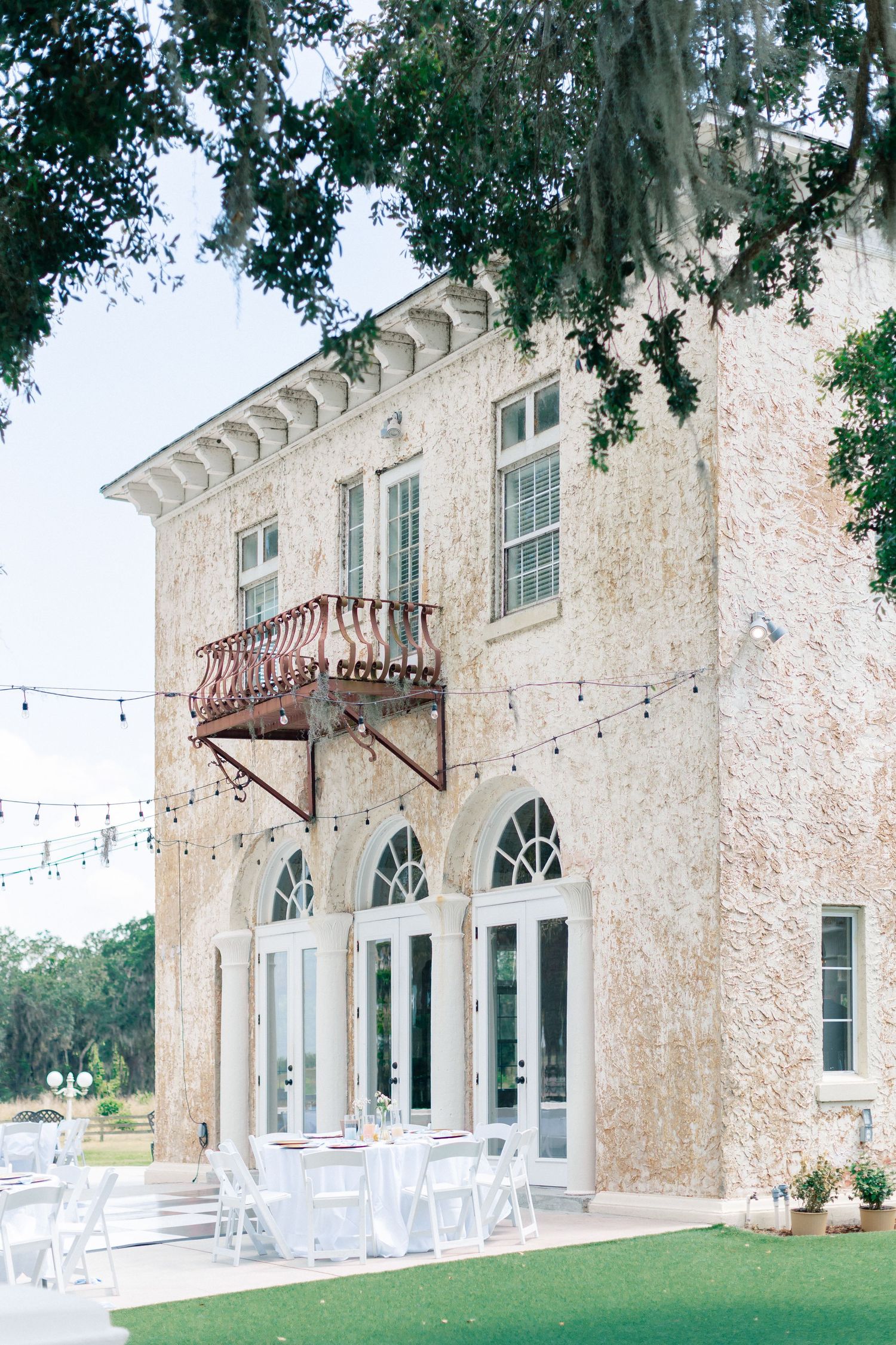 Elegant two-story historic venue with arched windows, balcony, string lights, and white chairs on manicured lawn, wedding at Bella Cosa