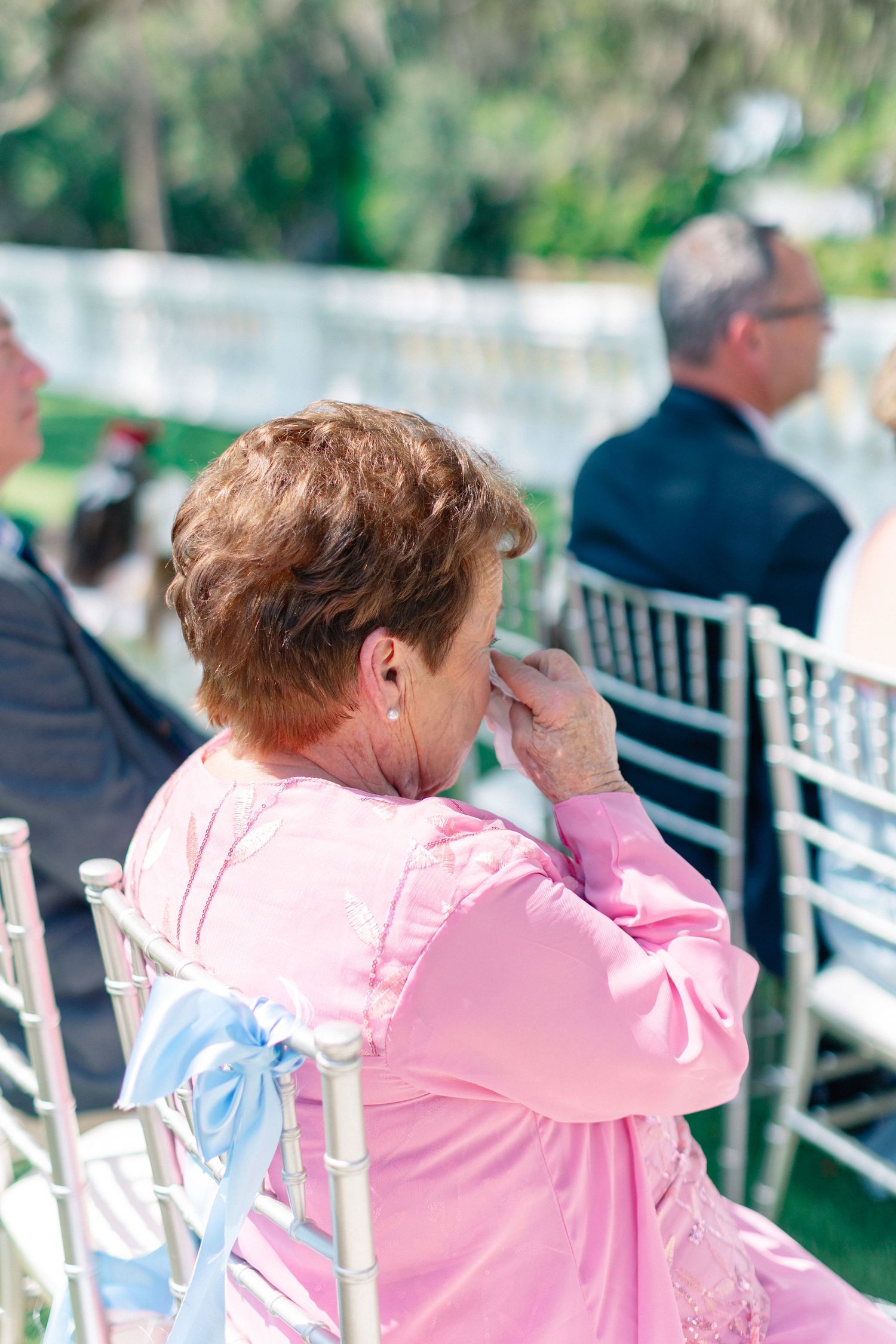 Elderly woman in pink dress wiping tears at outdoor wedding ceremony, seated on white chair, wedding at Bella Cosa