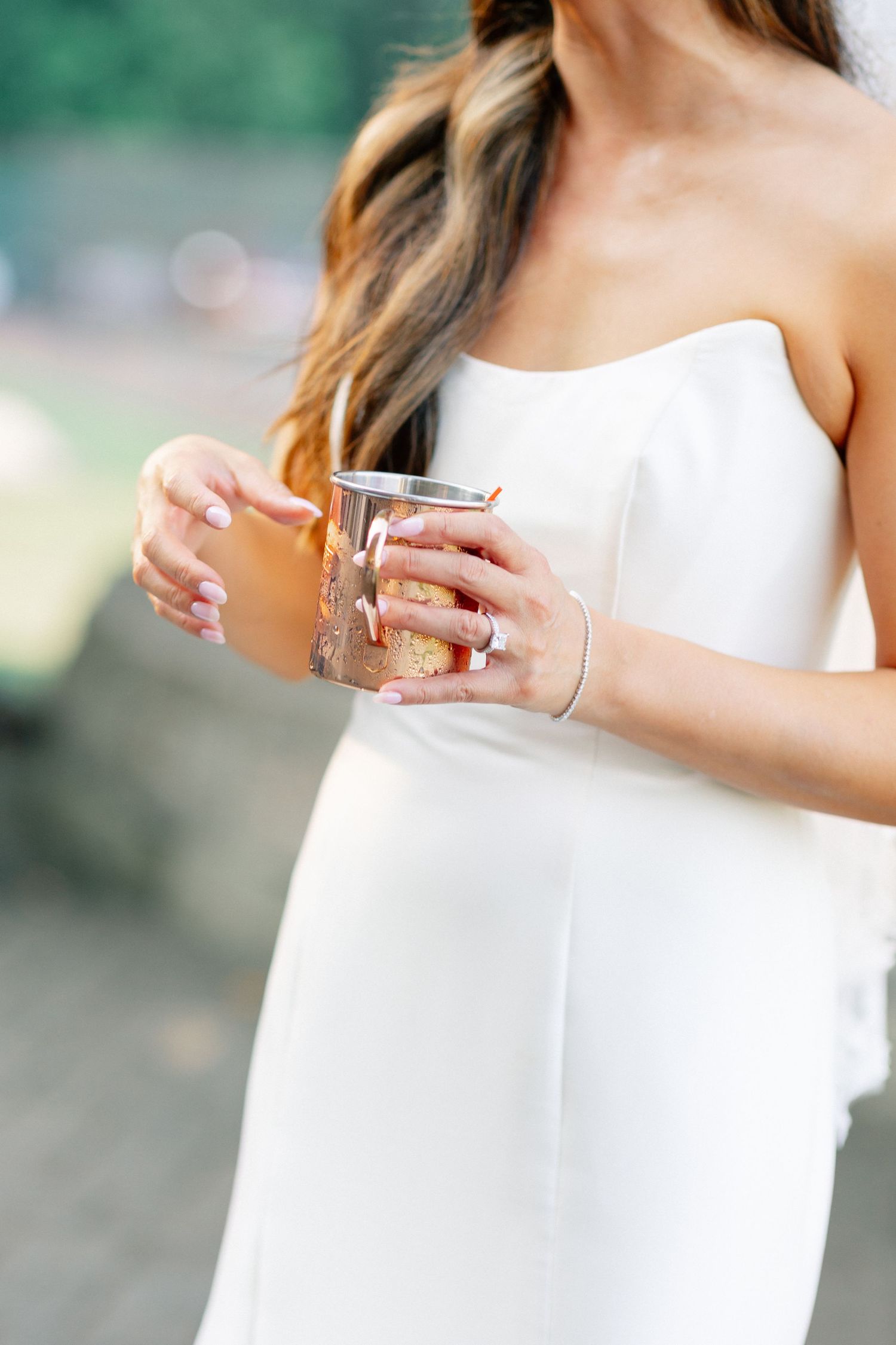 Bride in white strapless gown holding a glass drink, showcasing elegant manicure and bracelet at outdoor wedding.