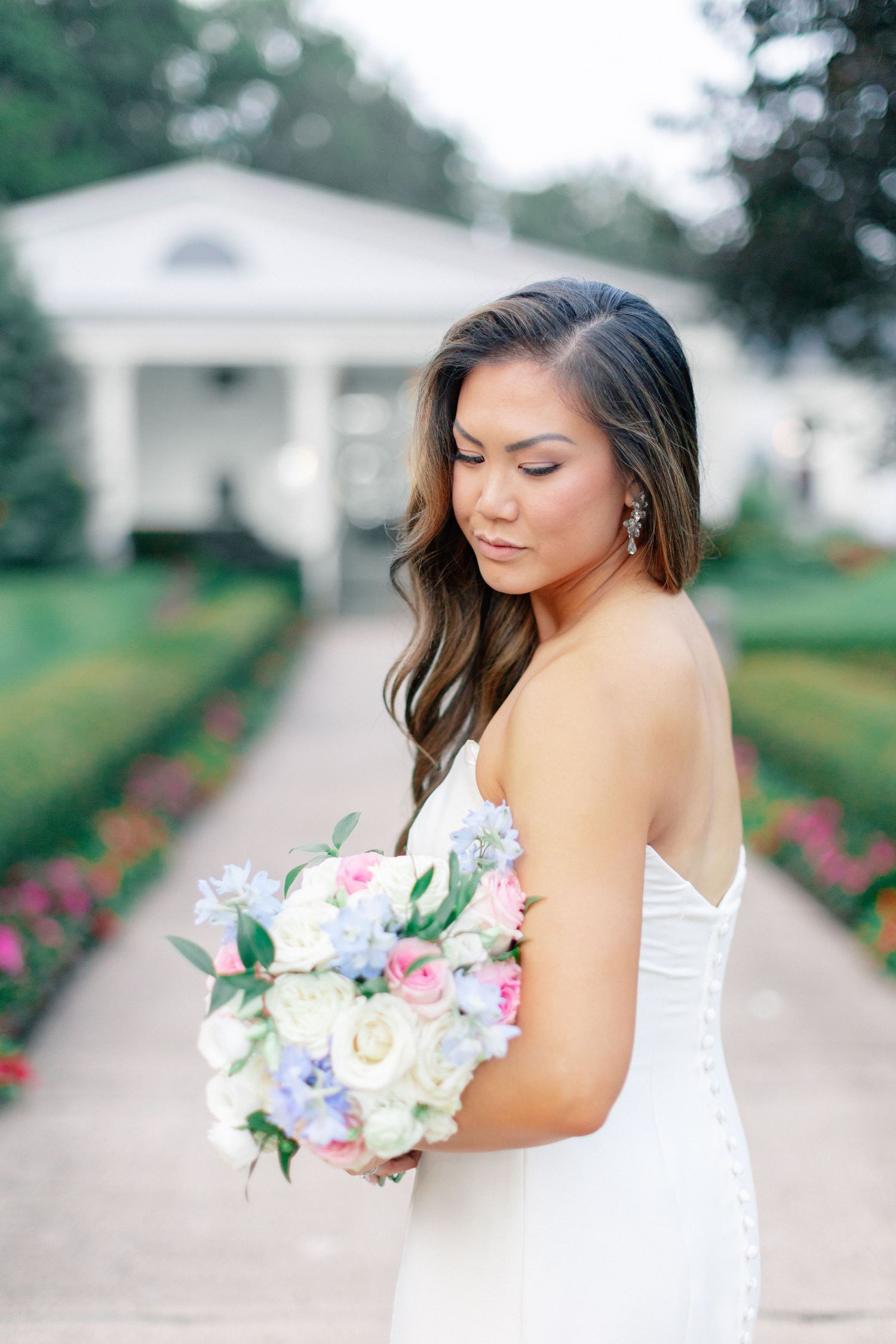 Bride in white strapless gown holding pastel bouquet of roses and blue flowers in a lush garden pathway.