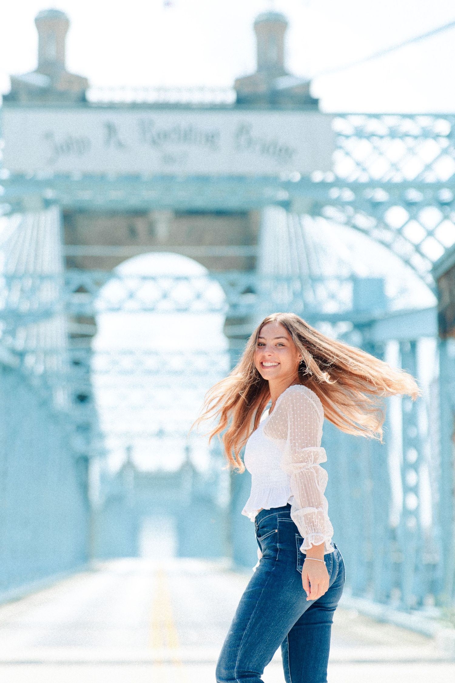 A smiling young woman with flowing hair poses on the John A. Roebling suspension bridge in a white top and jeans in Cincinnati Ohio