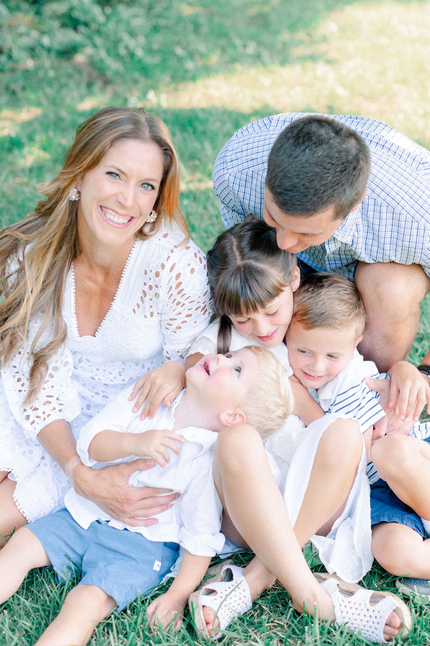 A happy family of five shares a warm embrace while sitting on green grass in a sunlit outdoor setting.