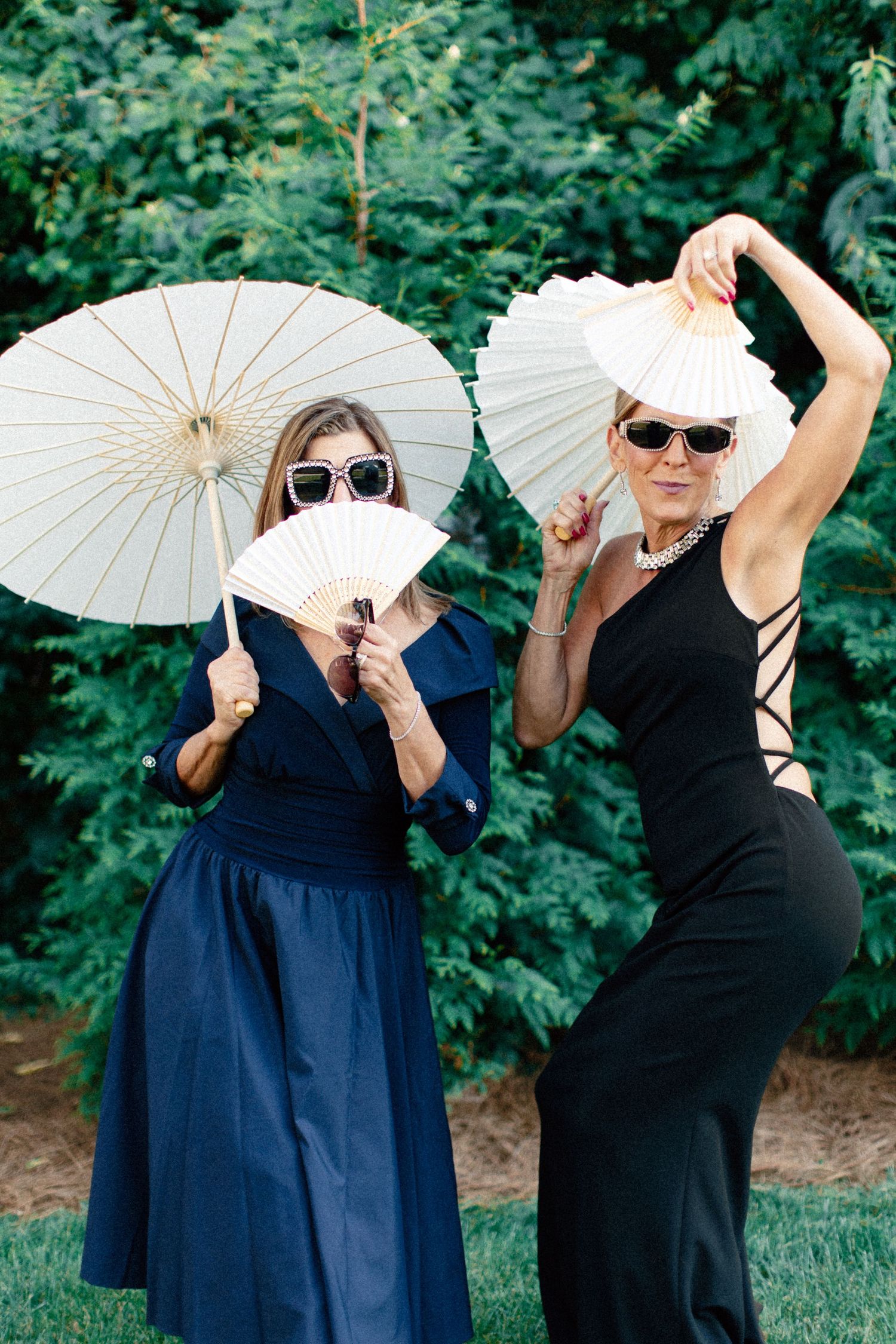 Two women in elegant dresses and sunglasses pose outdoors with white paper fans and parasol.