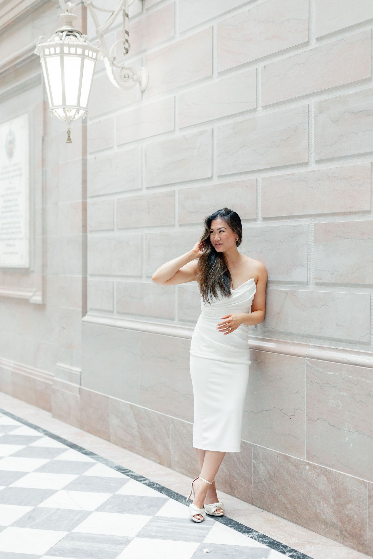 Bride in white strapless midi dress posing against stone wall with lantern.