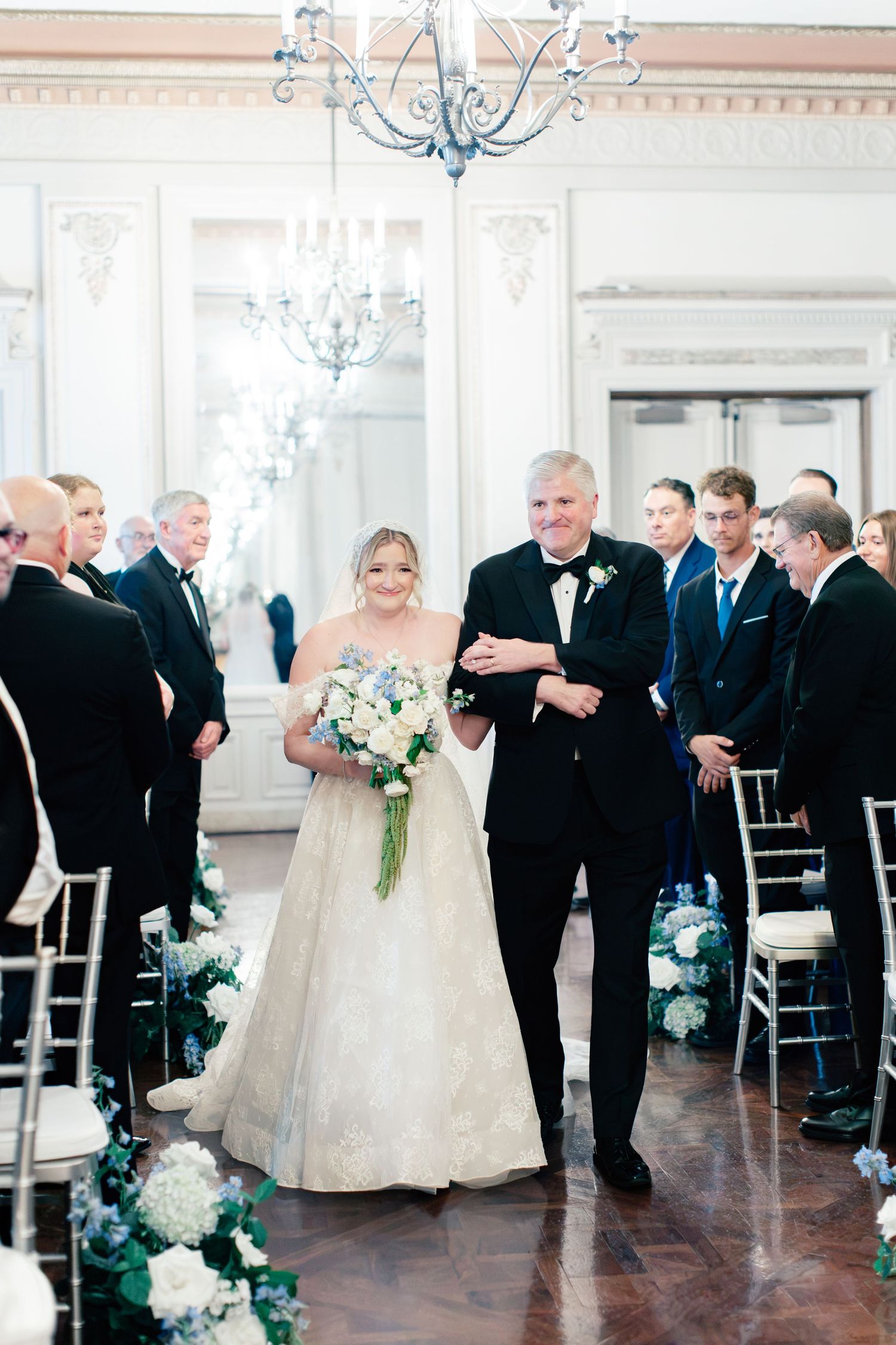 Bride in lace gown walks down aisle with escort in black tuxedo, holding blue and white bouquet in elegant ballroom.