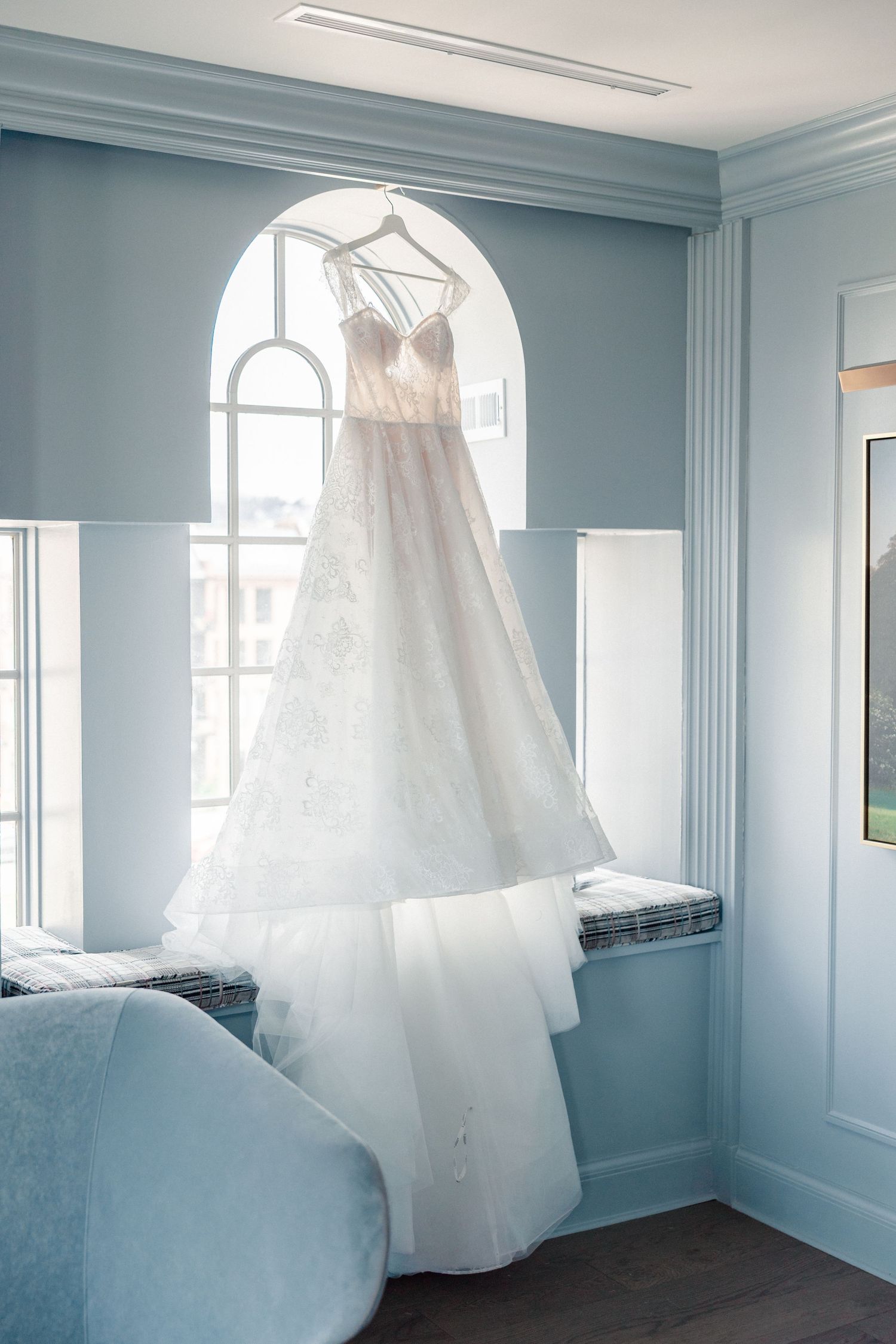 A elegant white lace wedding gown hanging in an arched window of a soft blue bridal suite.