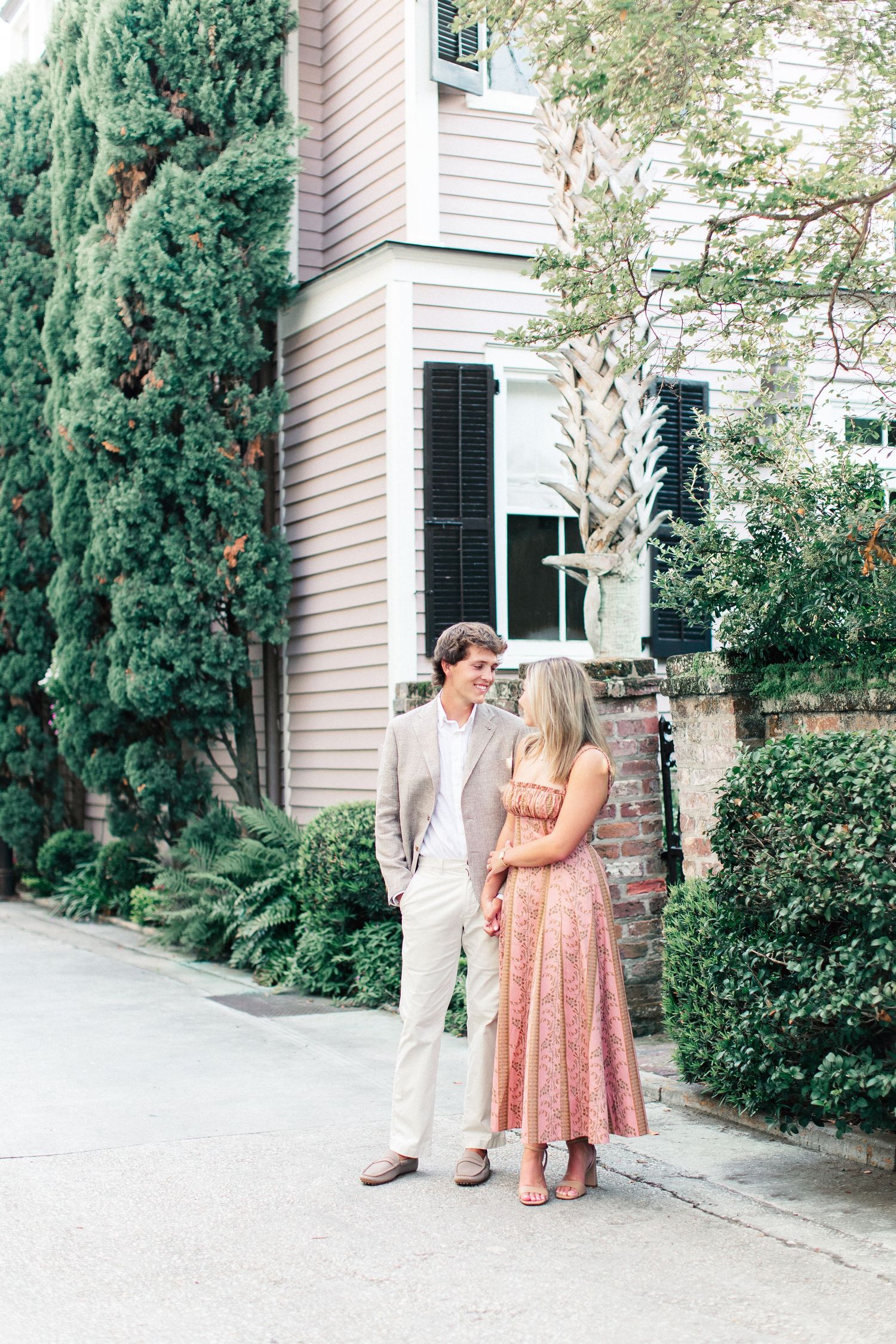 Couple standing together outside a pink historic home surrounded by lush greenery in Charleston, SC.