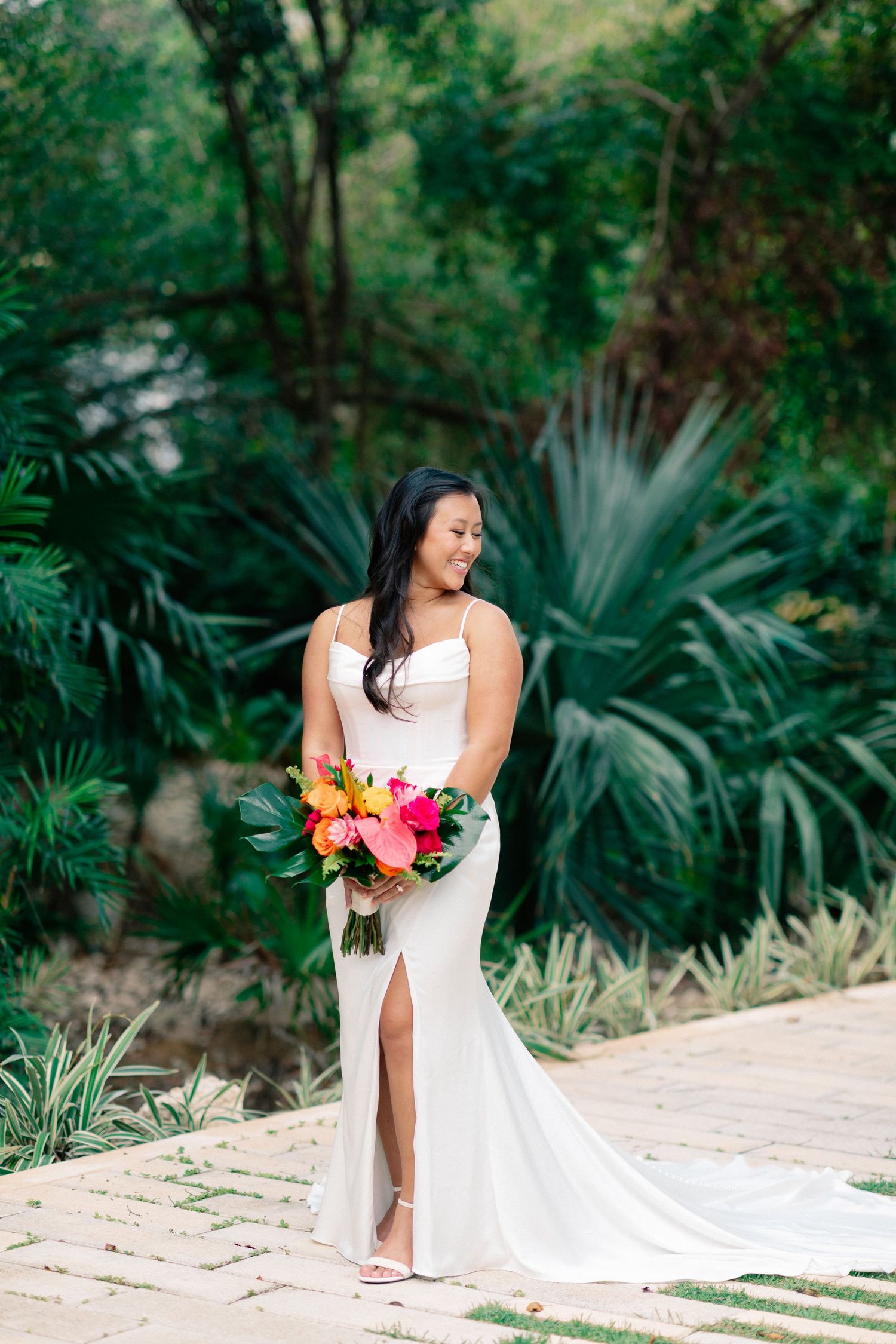 Bride in white gown holding tropical bouquet stands on stone path surrounded by lush green palms.