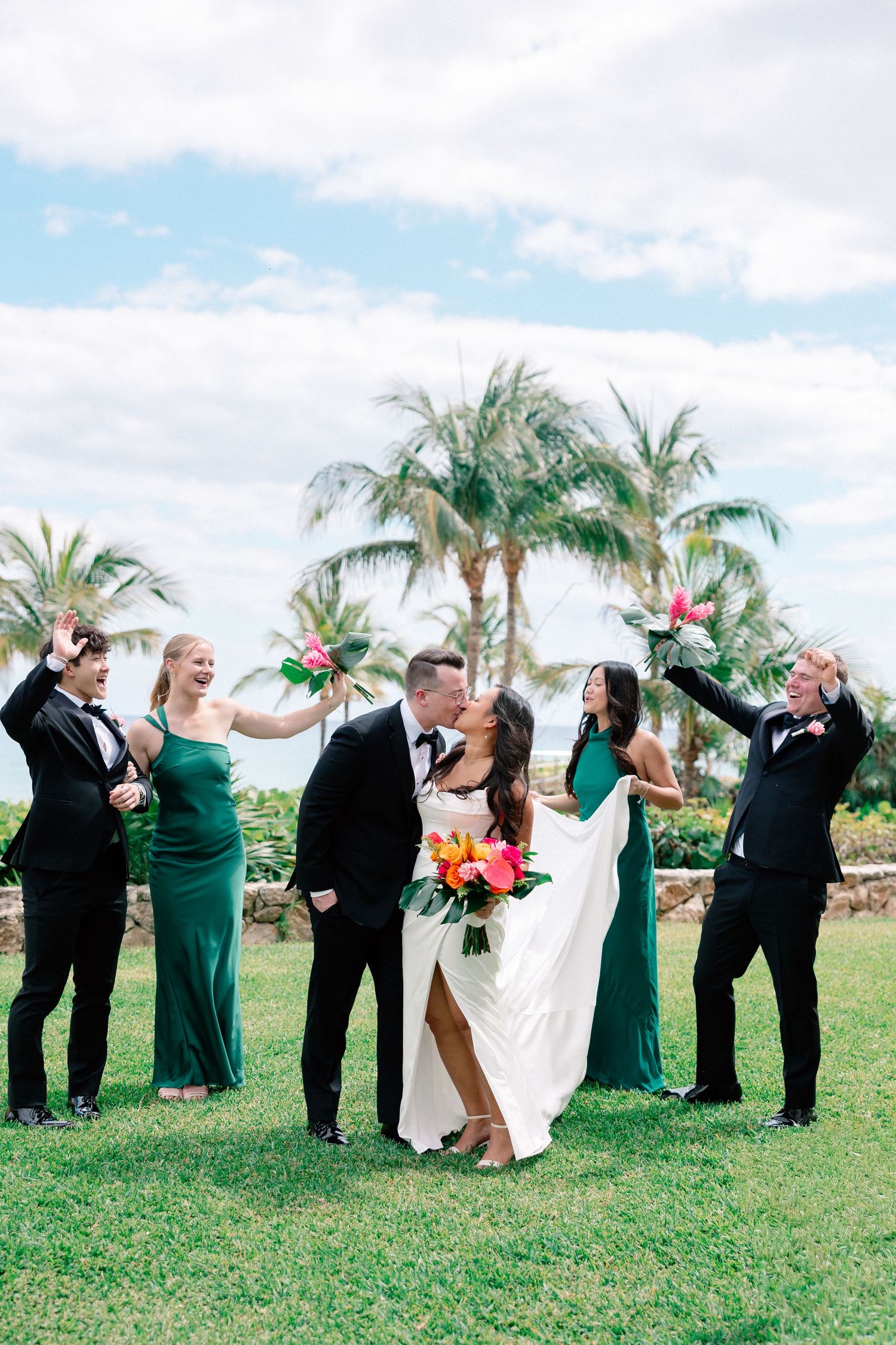 Bride and groom kiss surrounded by wedding party in emerald gowns and tuxedos on tropical lawn with palm trees.