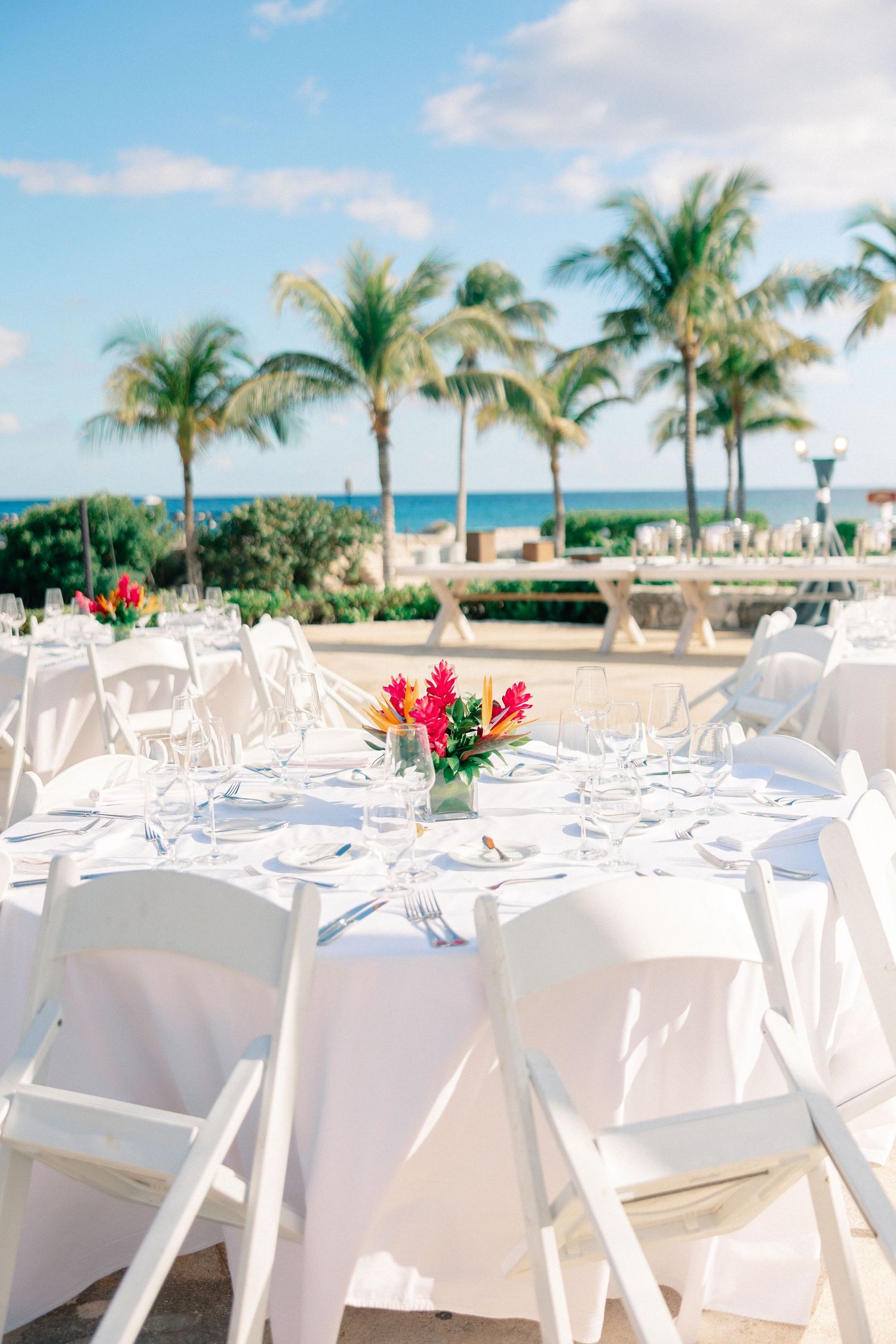 Elegant outdoor beachside wedding reception with white round tables, tropical floral centerpieces, and palm trees overlooking the ocean.