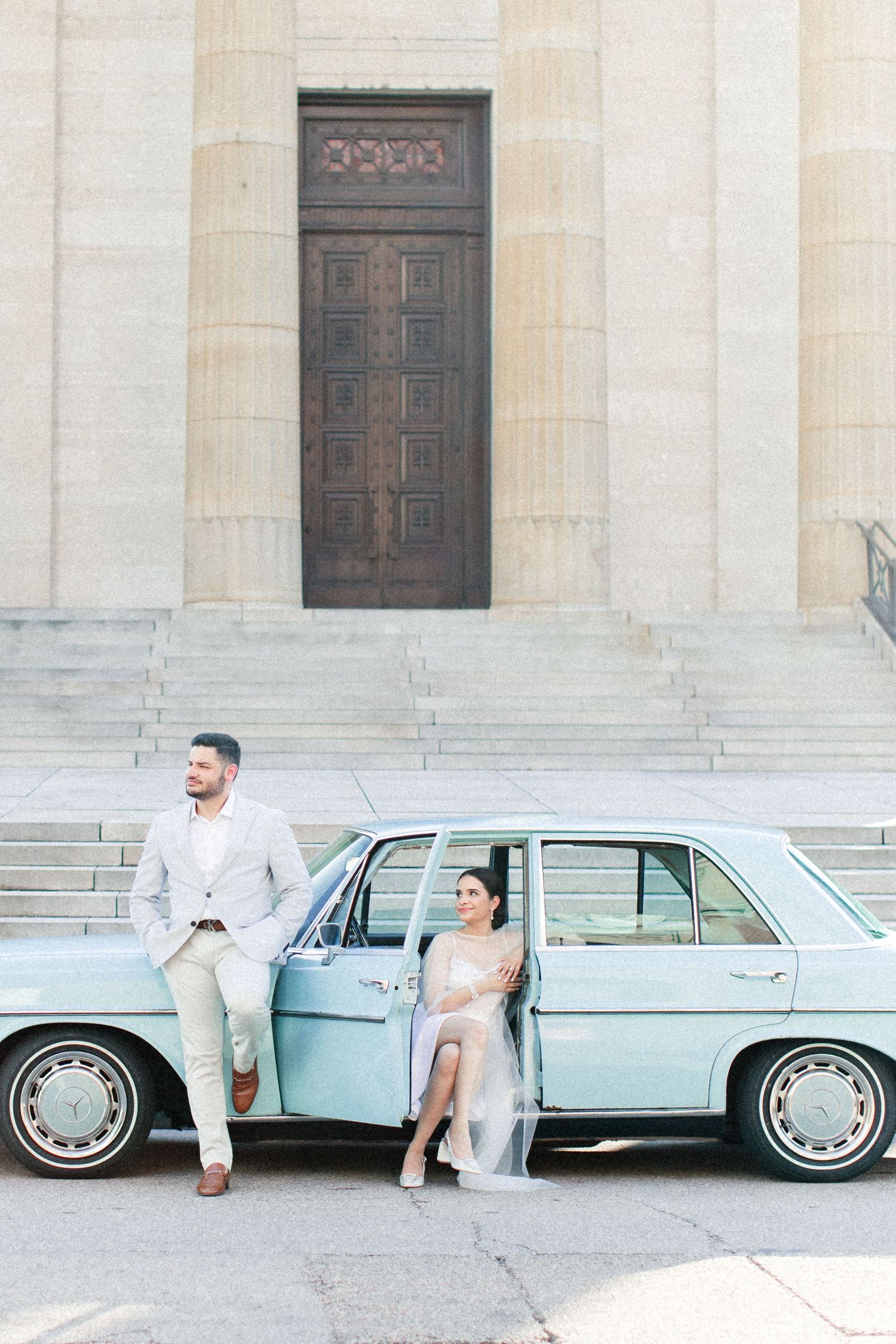 Bride and groom pose beside a vintage light blue car in front of a grand stone building with ornate wooden doors.