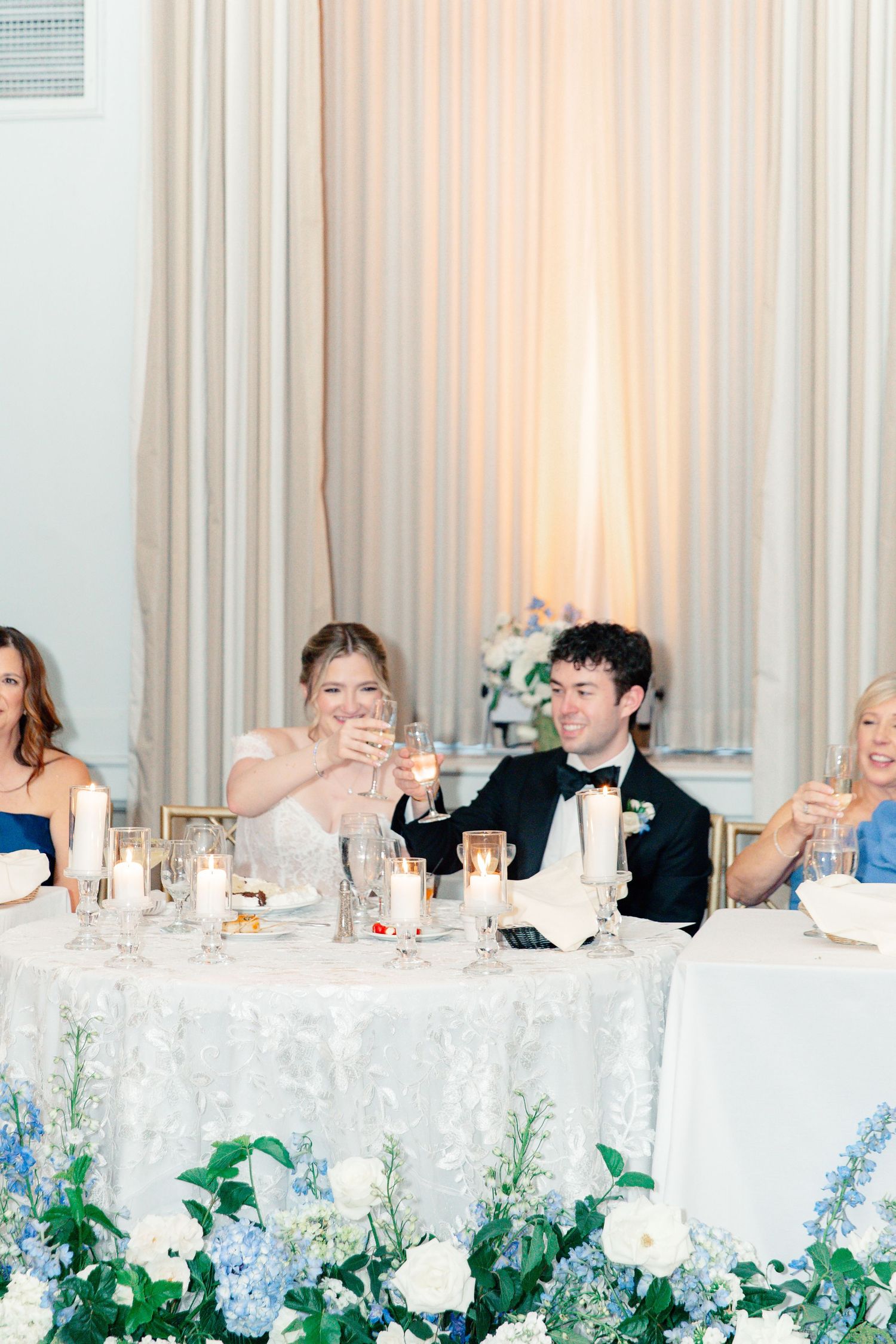 Bride and groom toast champagne at sweetheart table adorned with blue hydrangeas and white florals during wedding reception.
