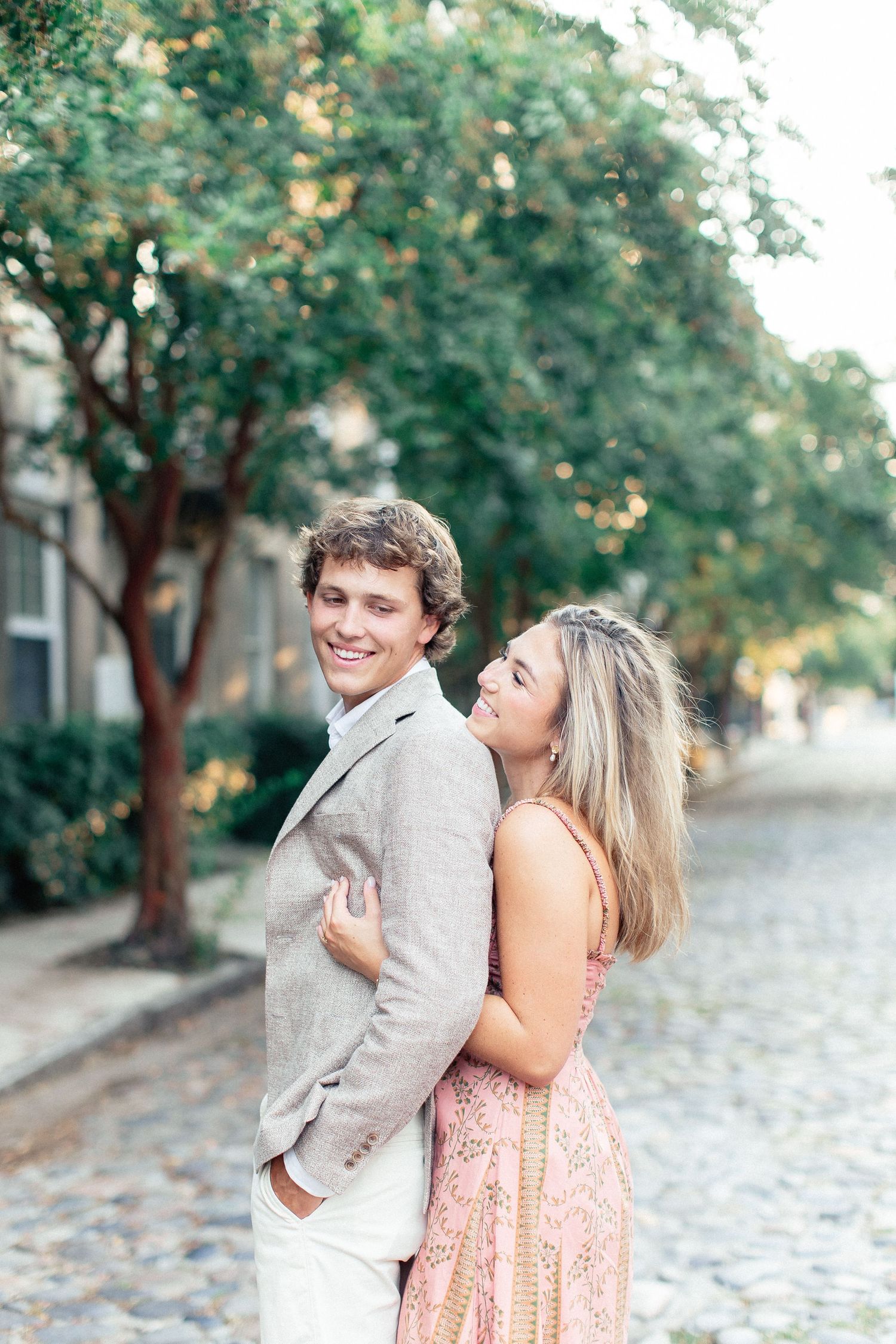 A smiling couple embraces on a cobblestone street lined with lush green trees, the woman in a pink dress hugging the man from behind.