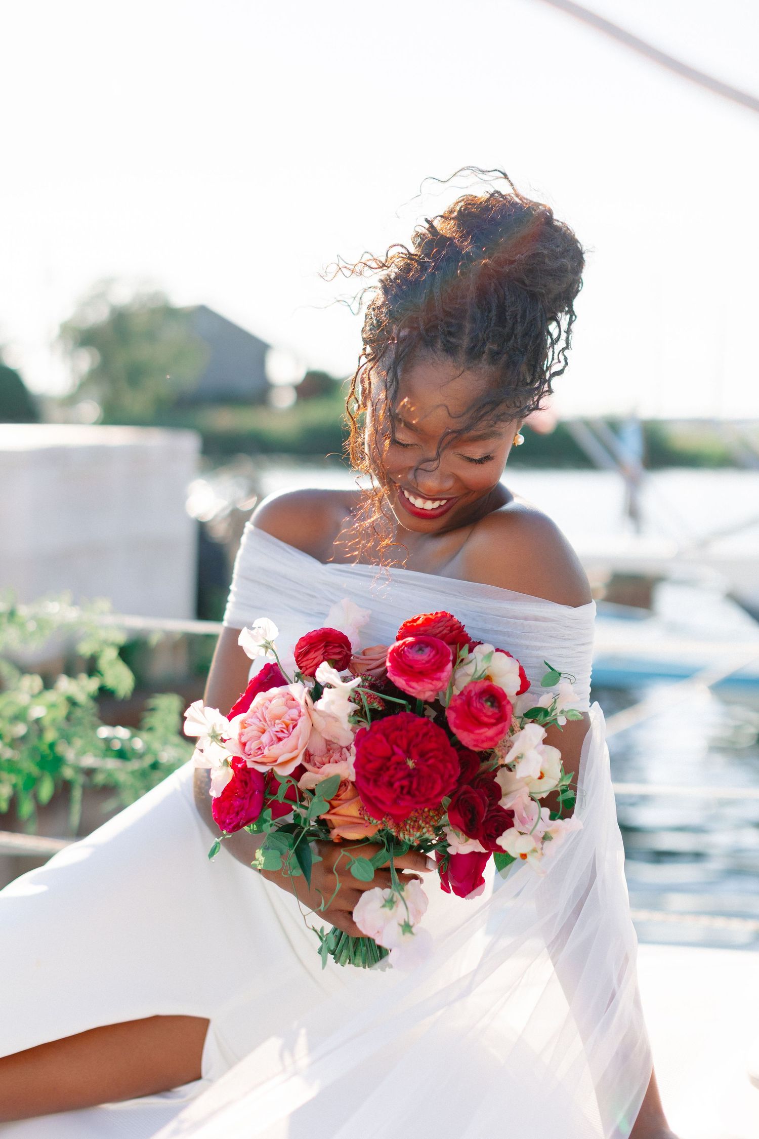 Smiling Black bride in white off-shoulder gown holding vibrant red and pink floral bouquet near marina.