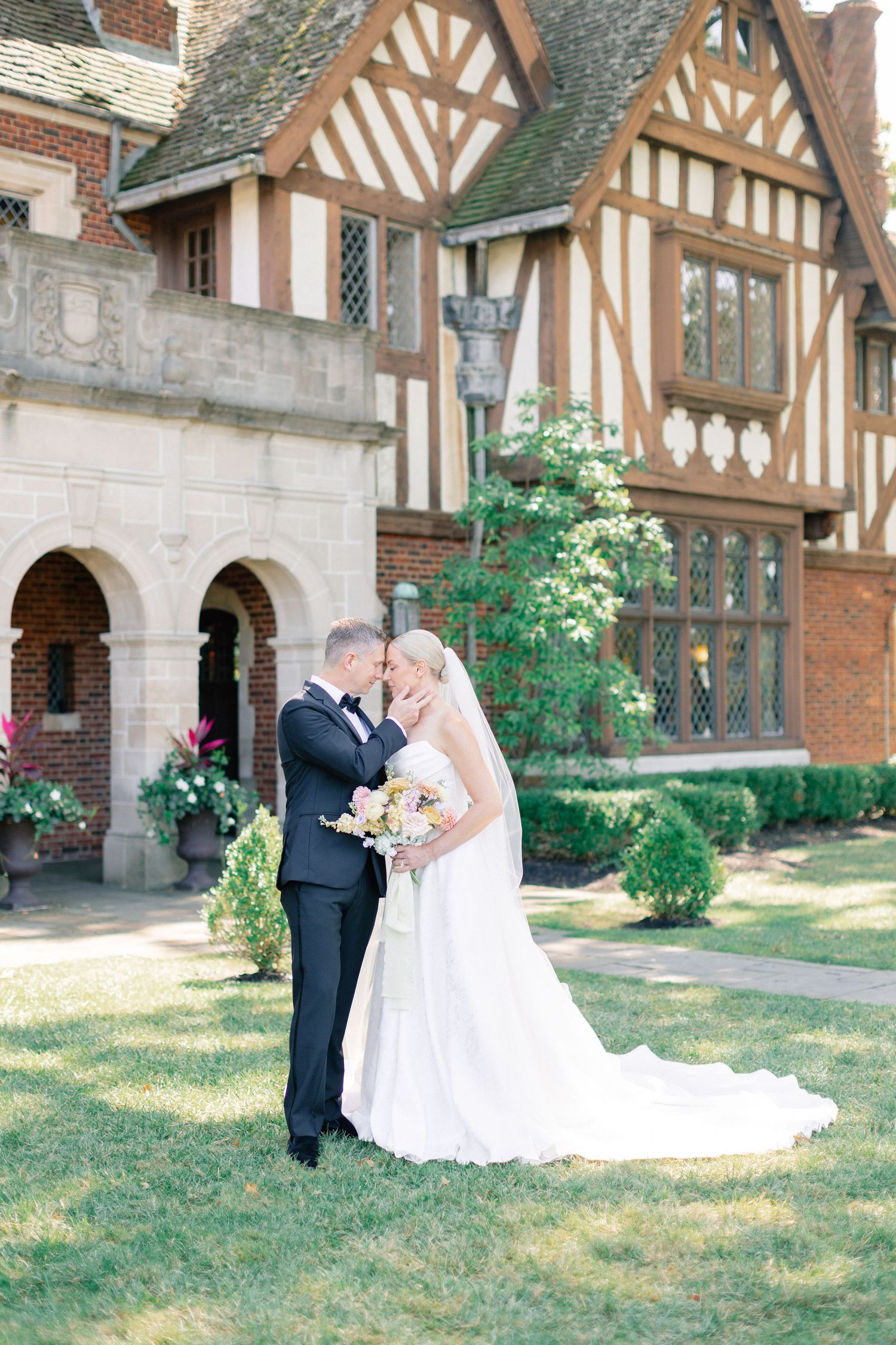 Bride and groom sharing romantic moment on lawn before Tudor-style brick manor wedding venue.
