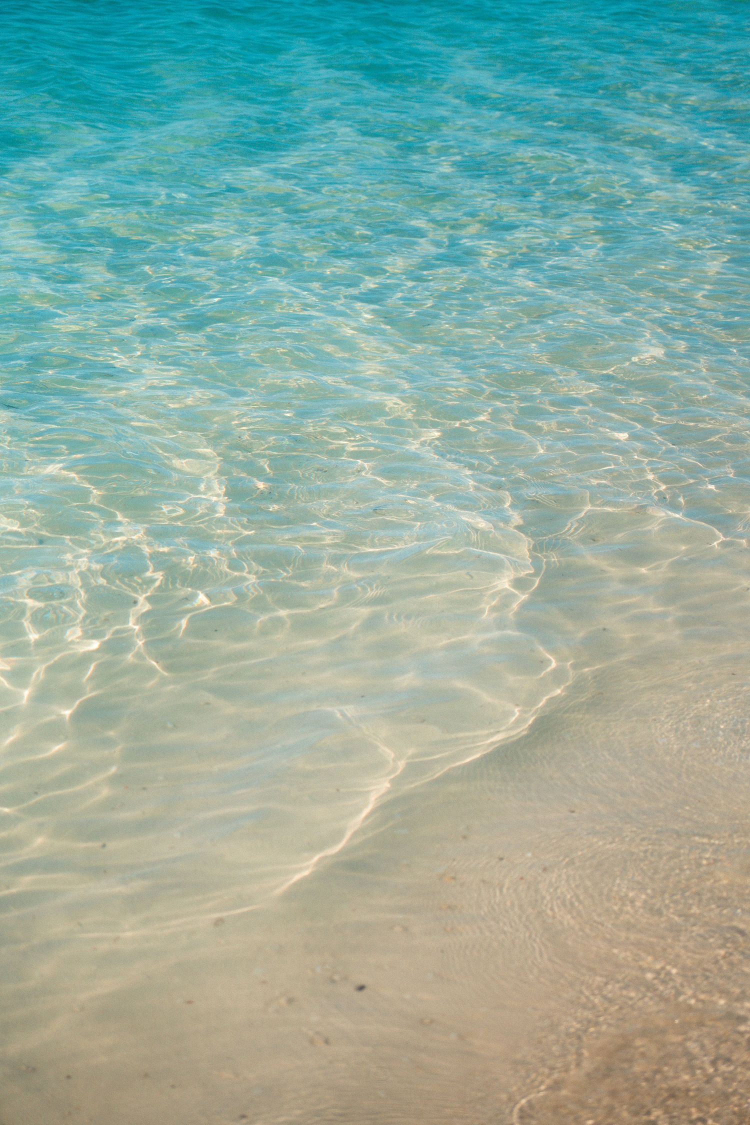 Crystal clear turquoise ocean water gently washing over white sandy beach shore in tropical paradise.