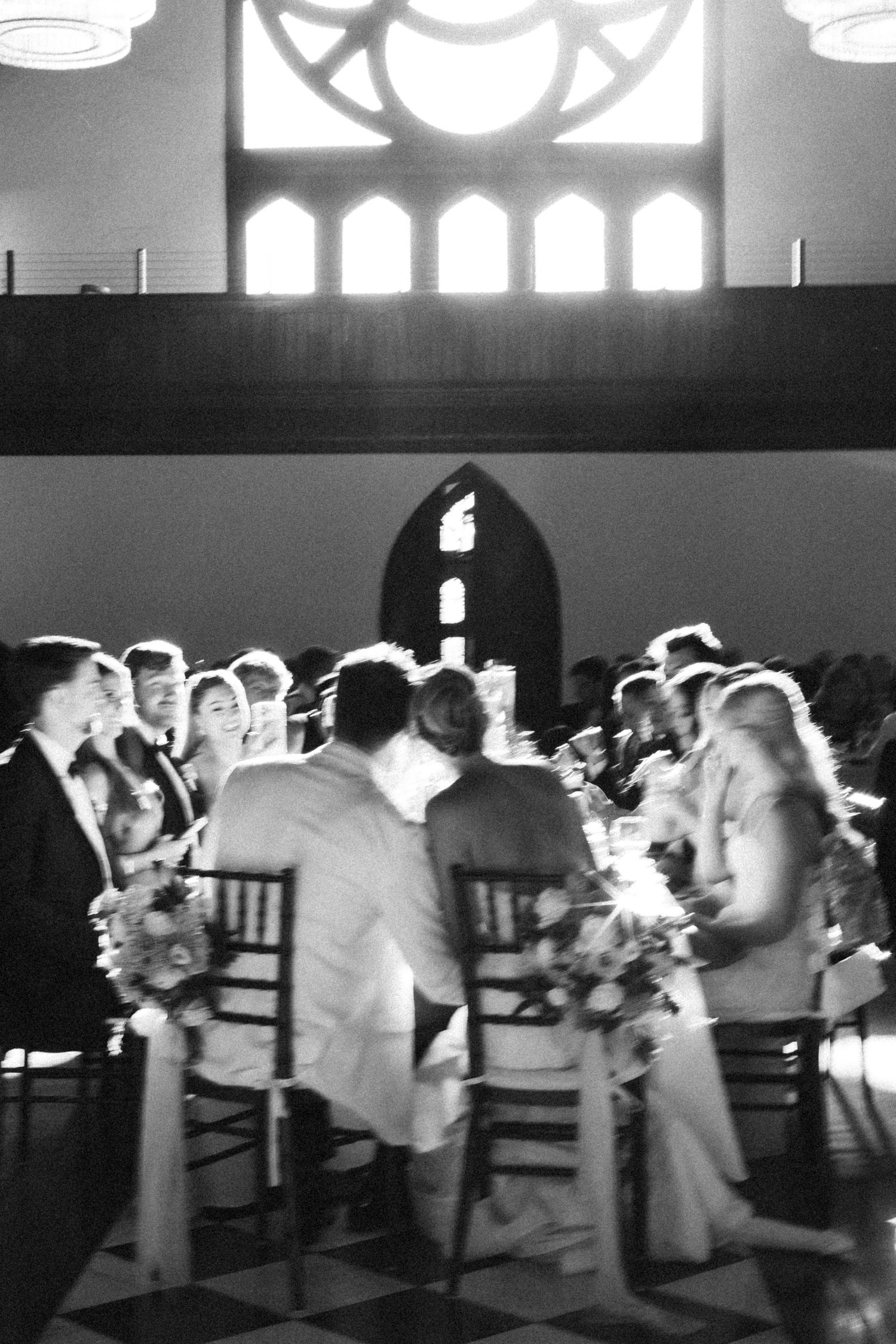 Black and white wedding reception photo inside a church venue with guests seated at tables adorned with floral centerpieces.