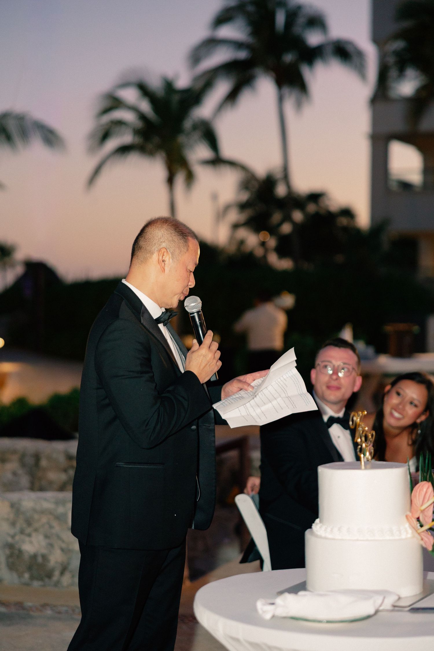 Man in tuxedo giving wedding toast with microphone and notes beside white cake at outdoor tropical reception.