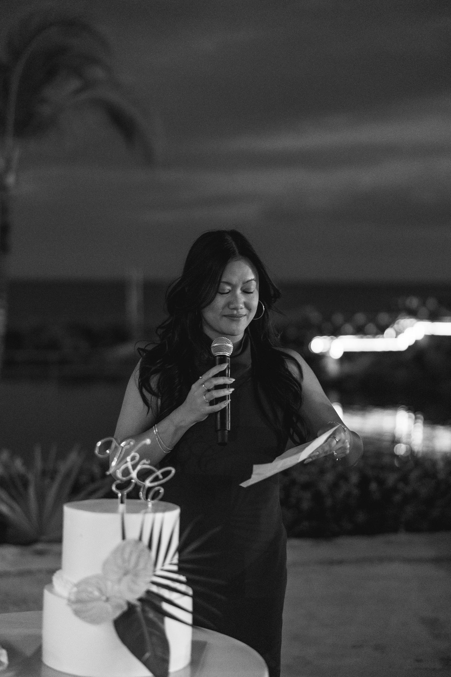 Woman in black dress speaking into microphone beside a wedding cake at an outdoor evening reception.