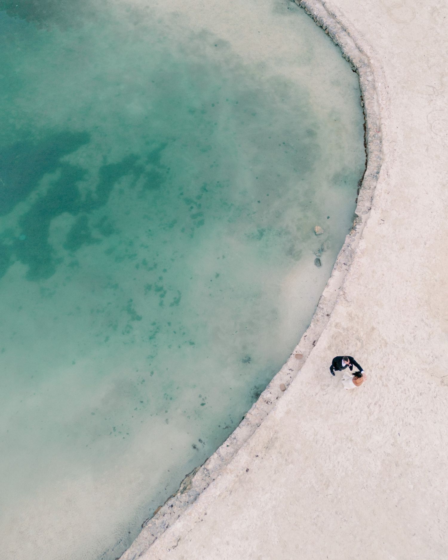 Aerial drone view of wedding couple embracing on white sandy shore beside stunning turquoise lagoon waters.
