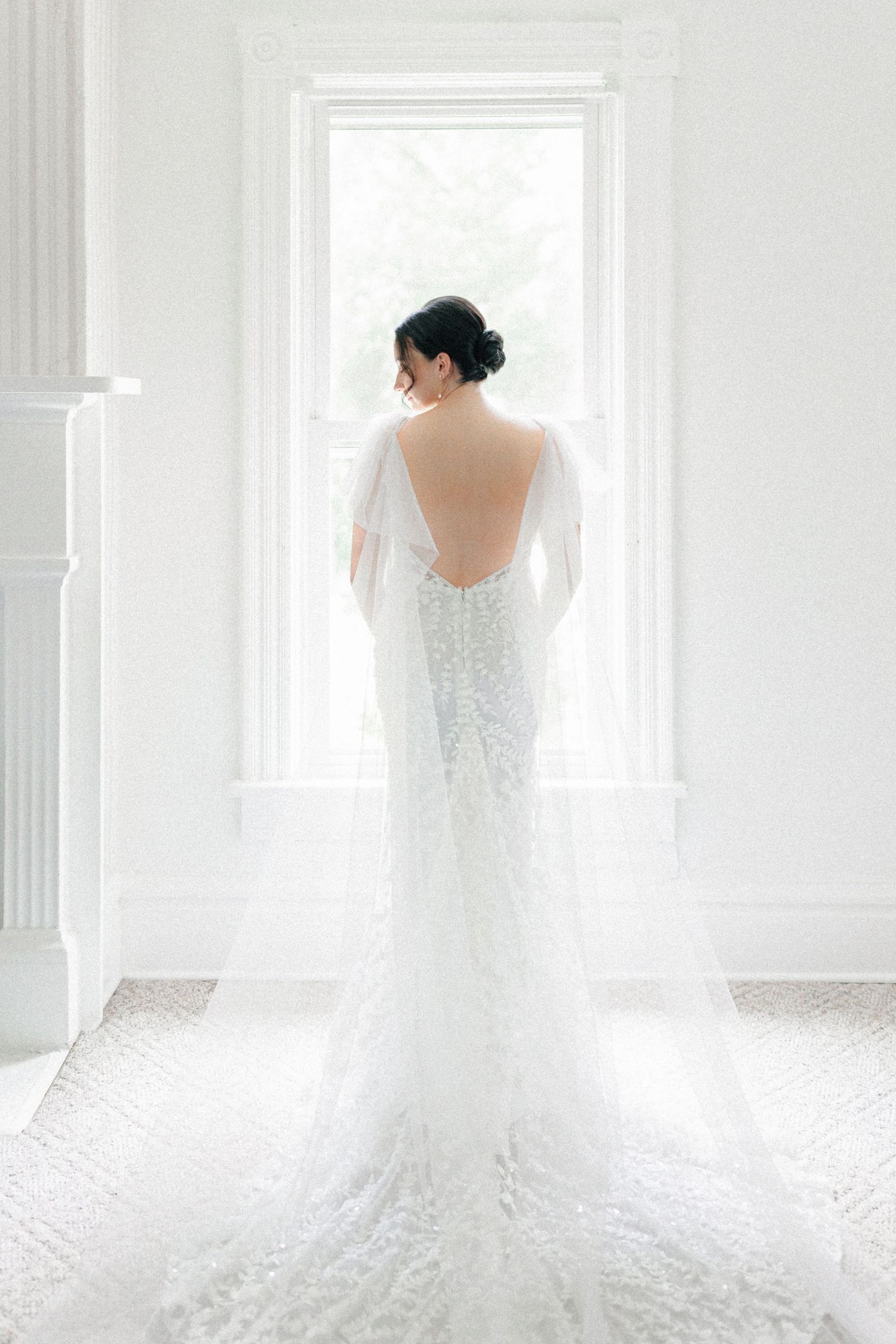 Elegant bride in a backless lace wedding gown with sheer cape train standing before a bright window.