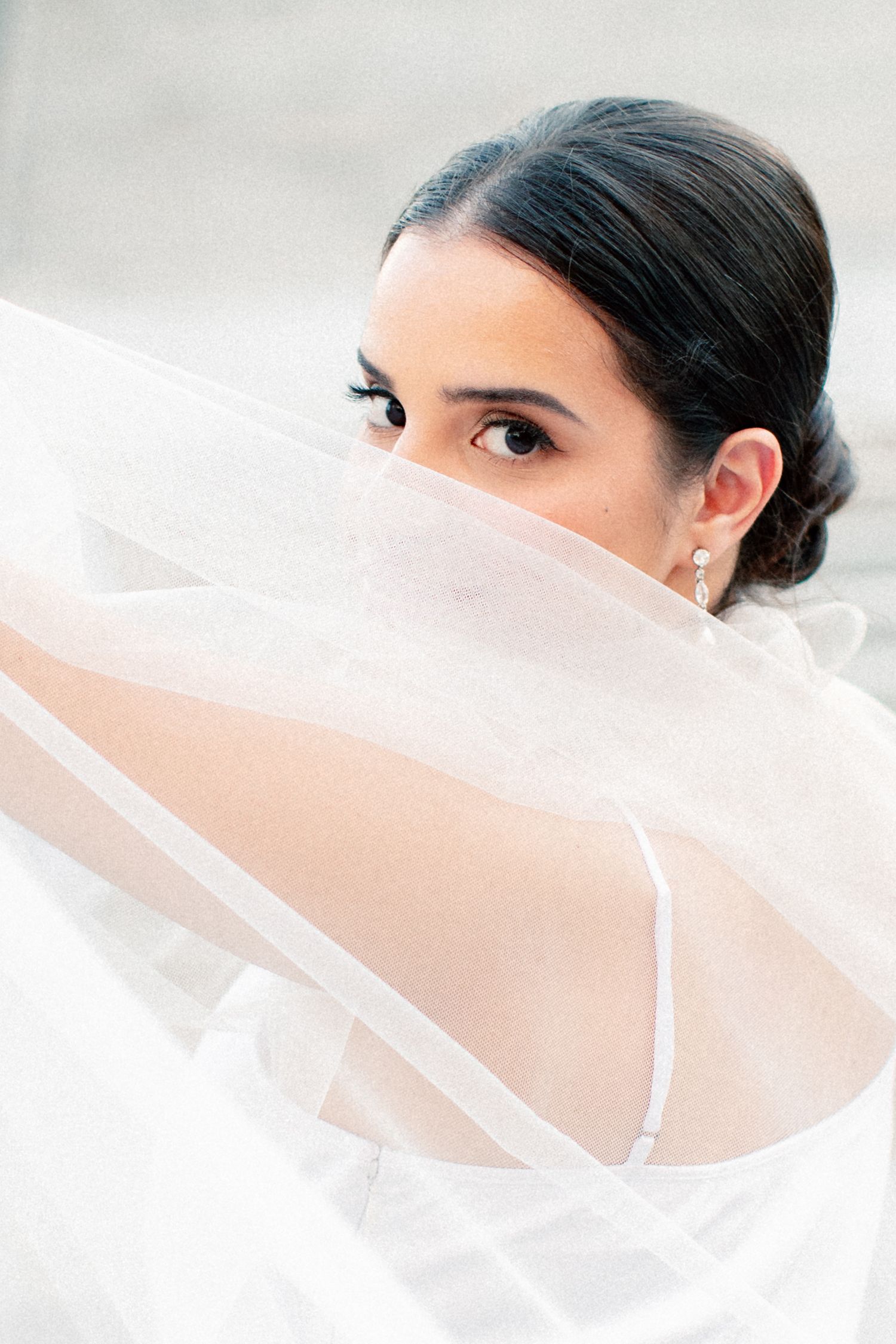 Bride with dark hair peers over flowing white veil, wearing elegant drop earrings in a soft, ethereal bridal portrait.