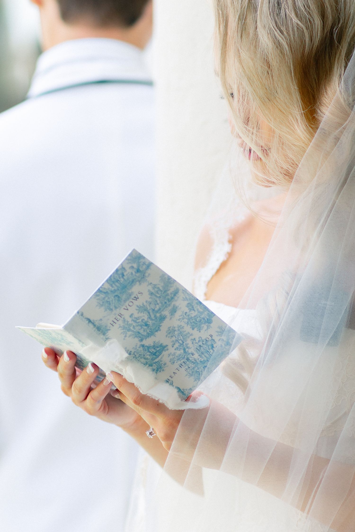 Bride in veil reads wedding vows from blue floral booklet, groom in white suit stands nearby, wedding at Bella Cosa