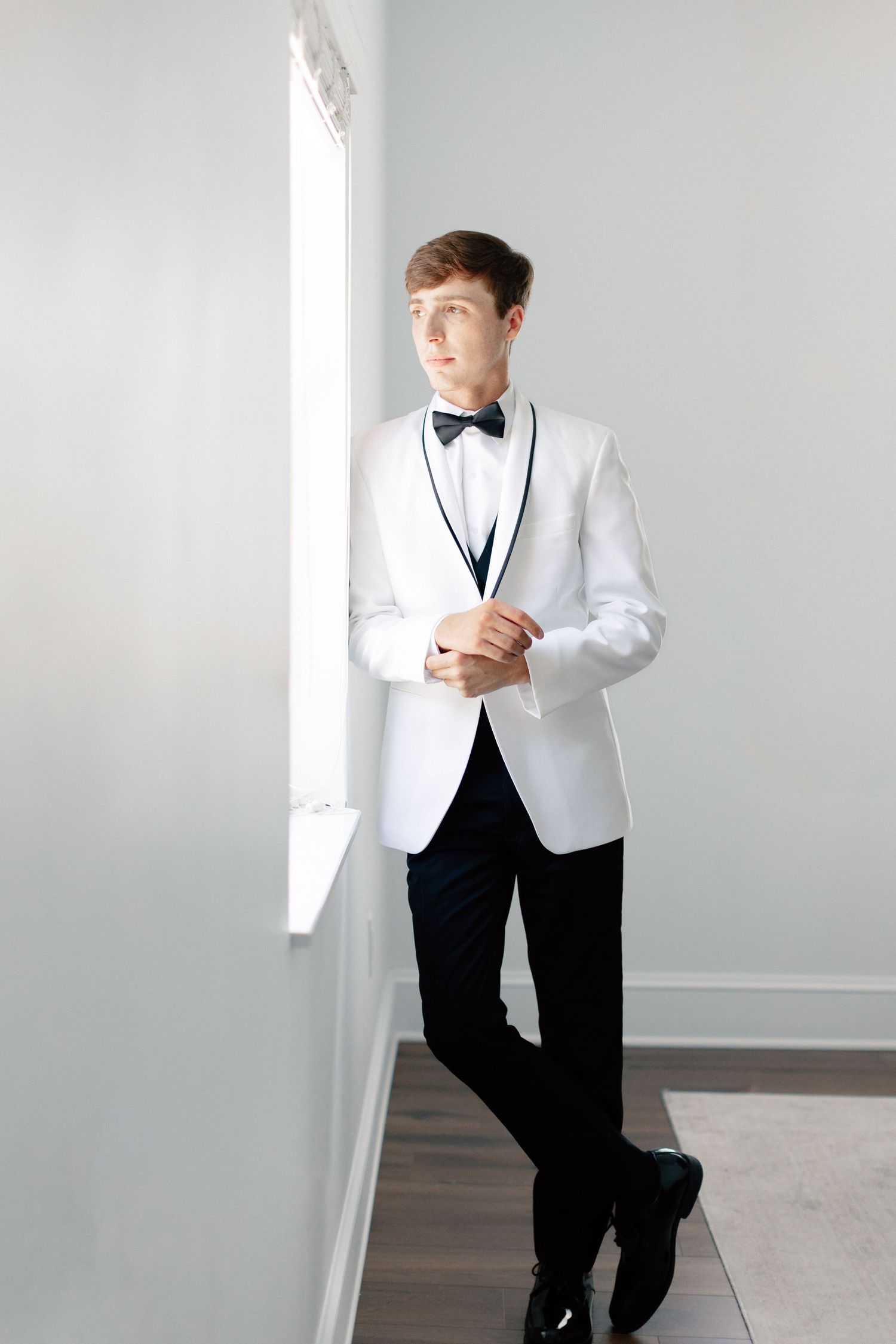 Young man in white tuxedo jacket with black bow tie and pants stands by window in elegant formal attire, wedding at Bella Cosa