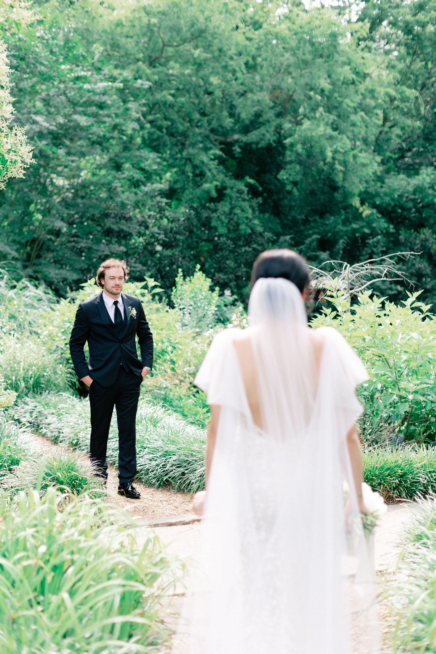 Bride in white gown and veil faces groom in black suit on a garden path surrounded by lush greenery.