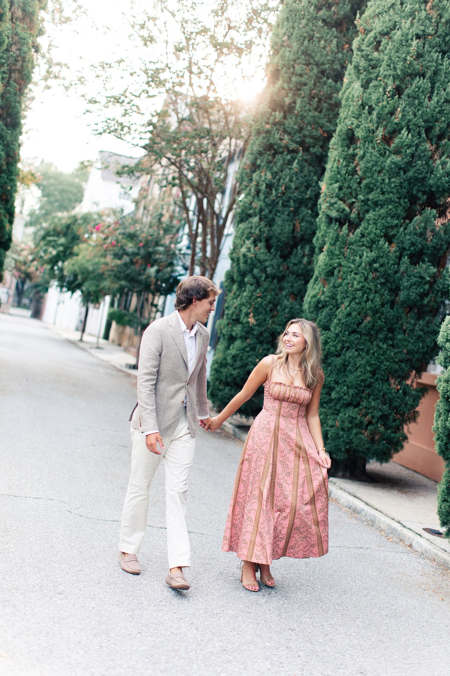 Couple holding hands walking down a tree-lined street, woman in pink floral dress and man in beige suit.