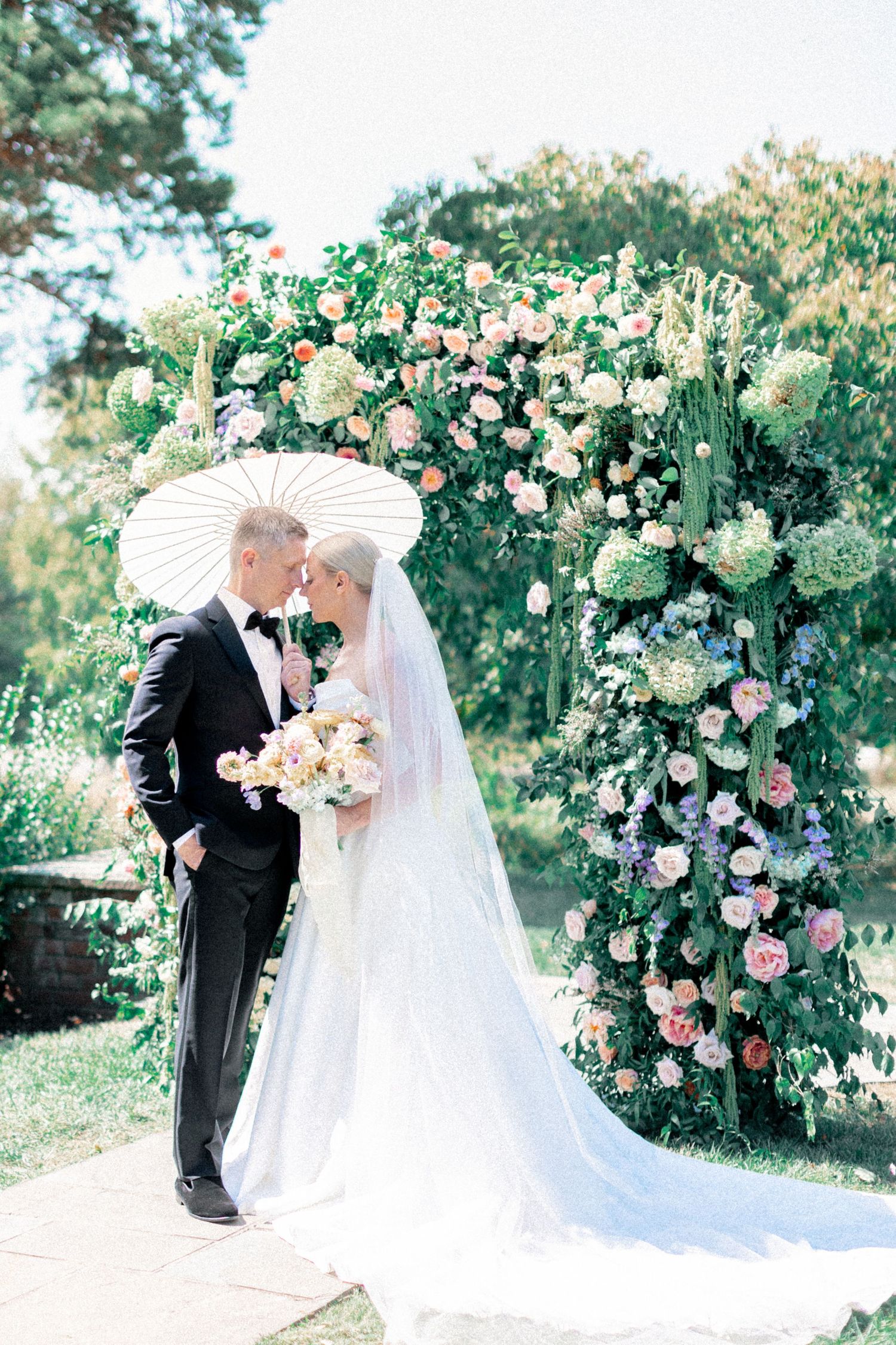 Bride and groom share a romantic moment under a parasol before a lush floral arch at an outdoor garden wedding.