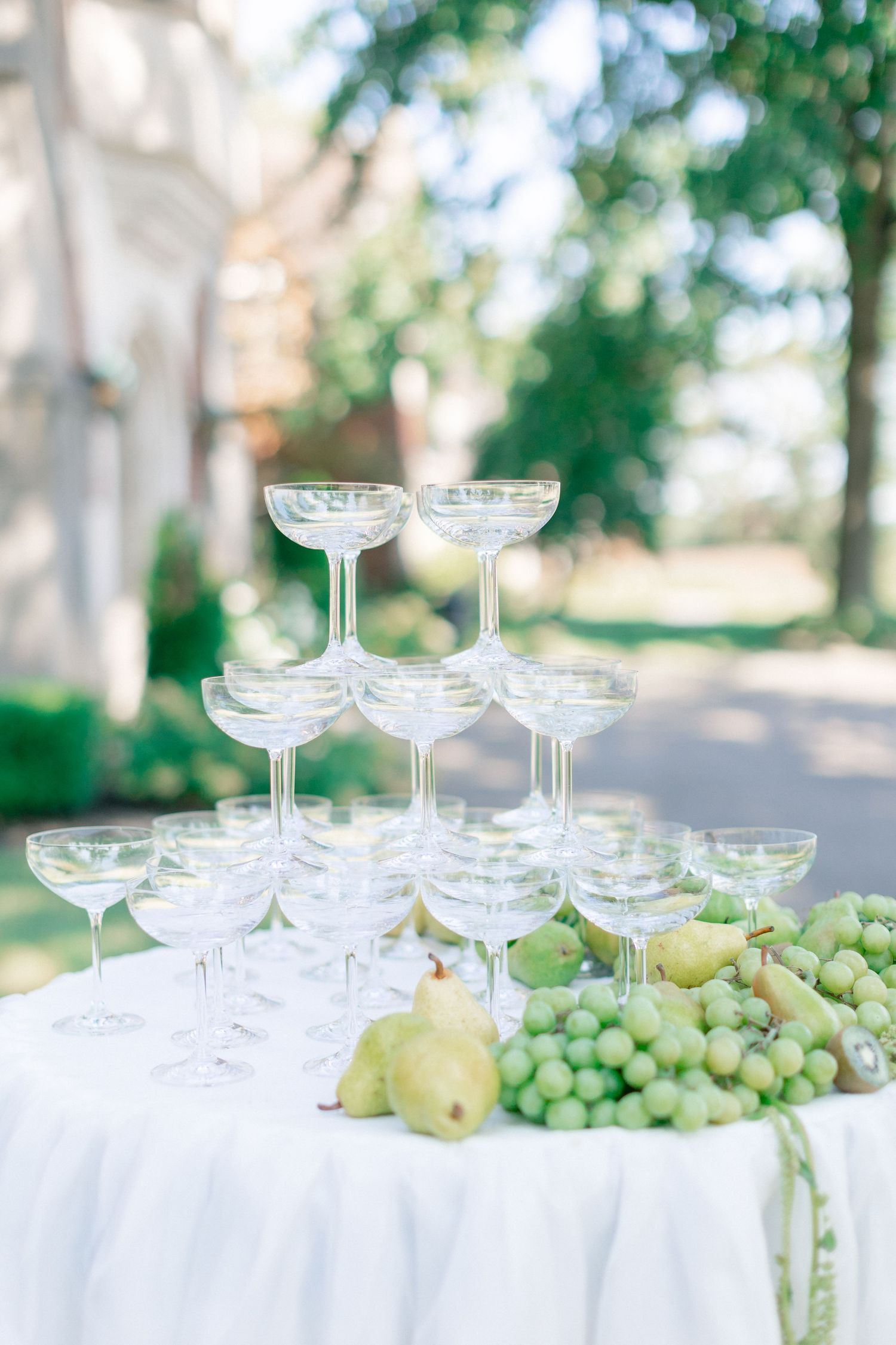 Champagne tower of coupe glasses surrounded by fresh green grapes and pears on a white draped table outdoors.