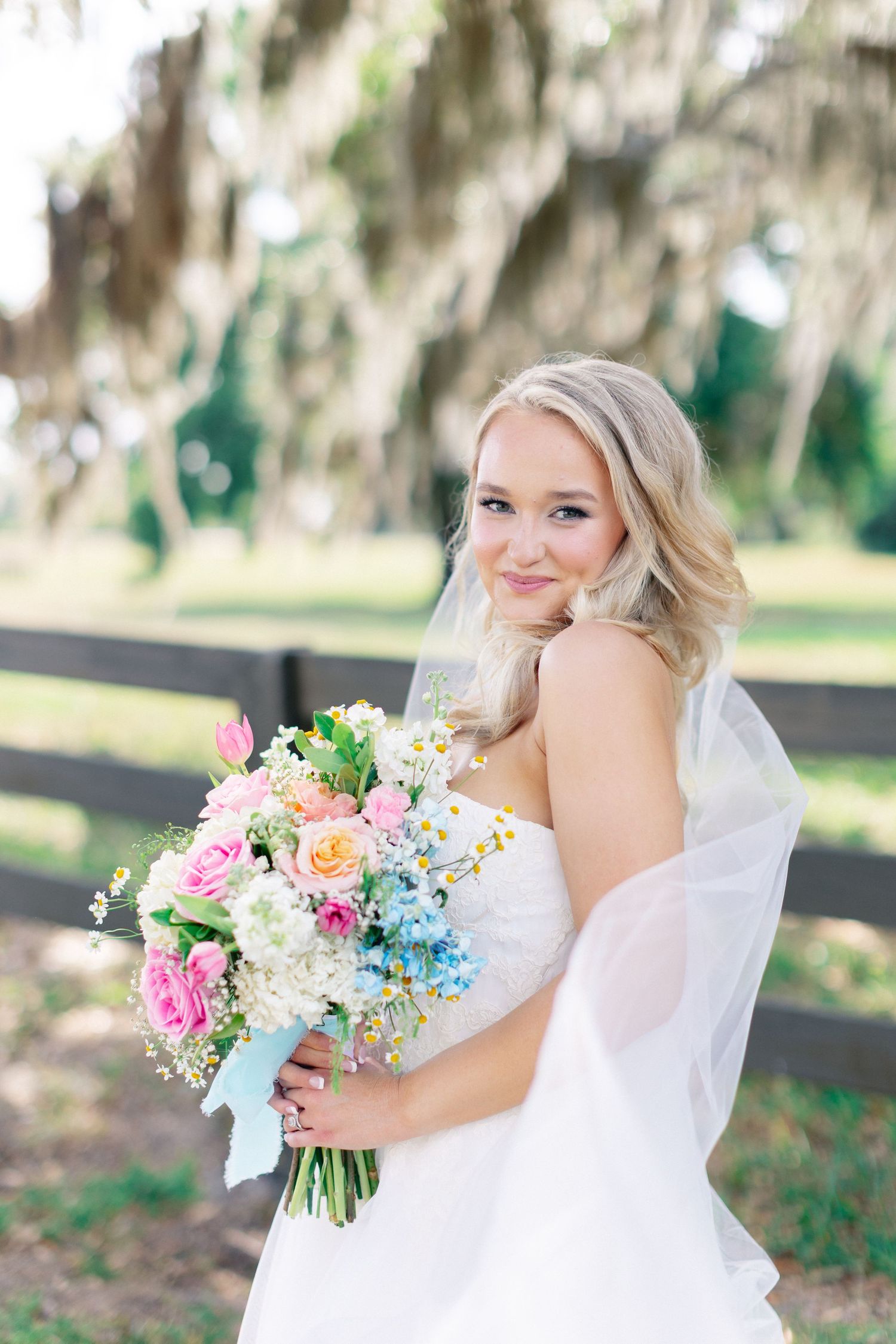 Smiling bride in white gown holds colorful spring bouquet with pink, blue, and peach flowers outdoors, wedding at Bella Cosa