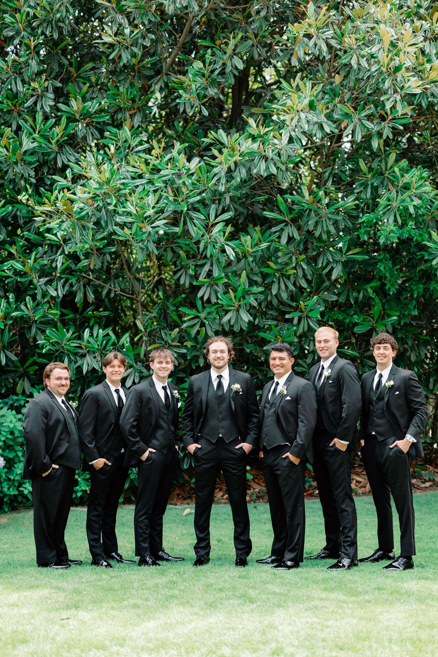 Groom and groomsmen in black suits posing outdoors in front of lush green trees on a wedding day.