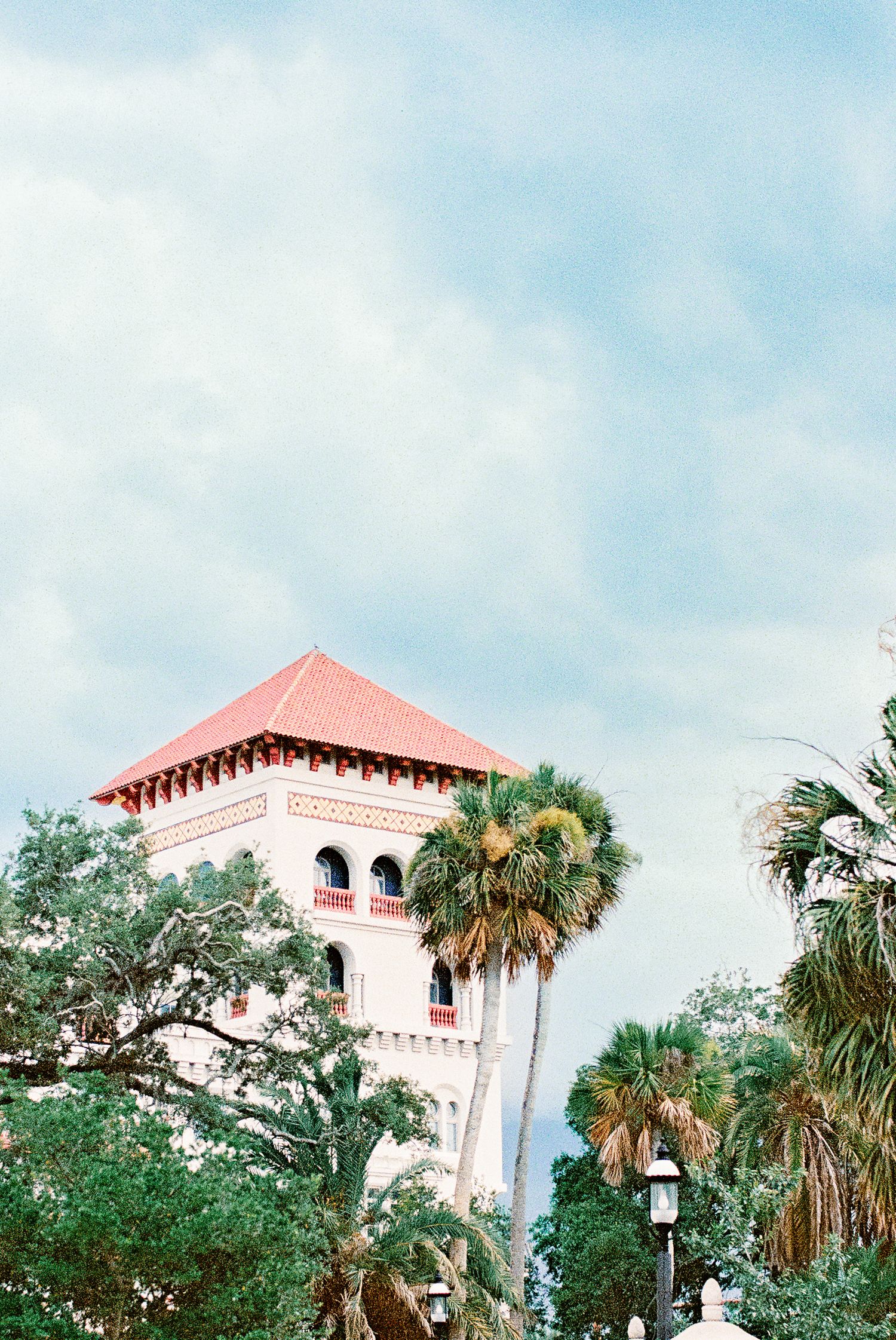 Historic pink tower with red hip roof rises above lush palm trees against a blue cloudy sky in a tropical setting.