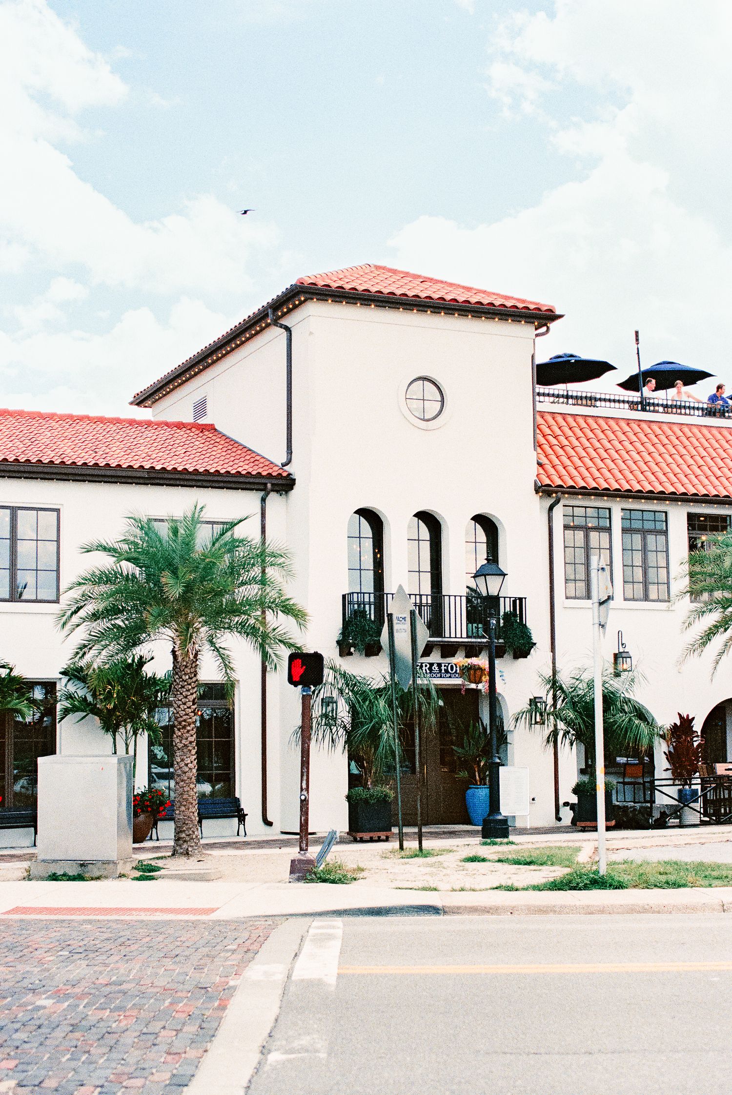 Spanish-style white building with red tile roof, palm trees, and cobblestone street corner in a sunny Florida town.