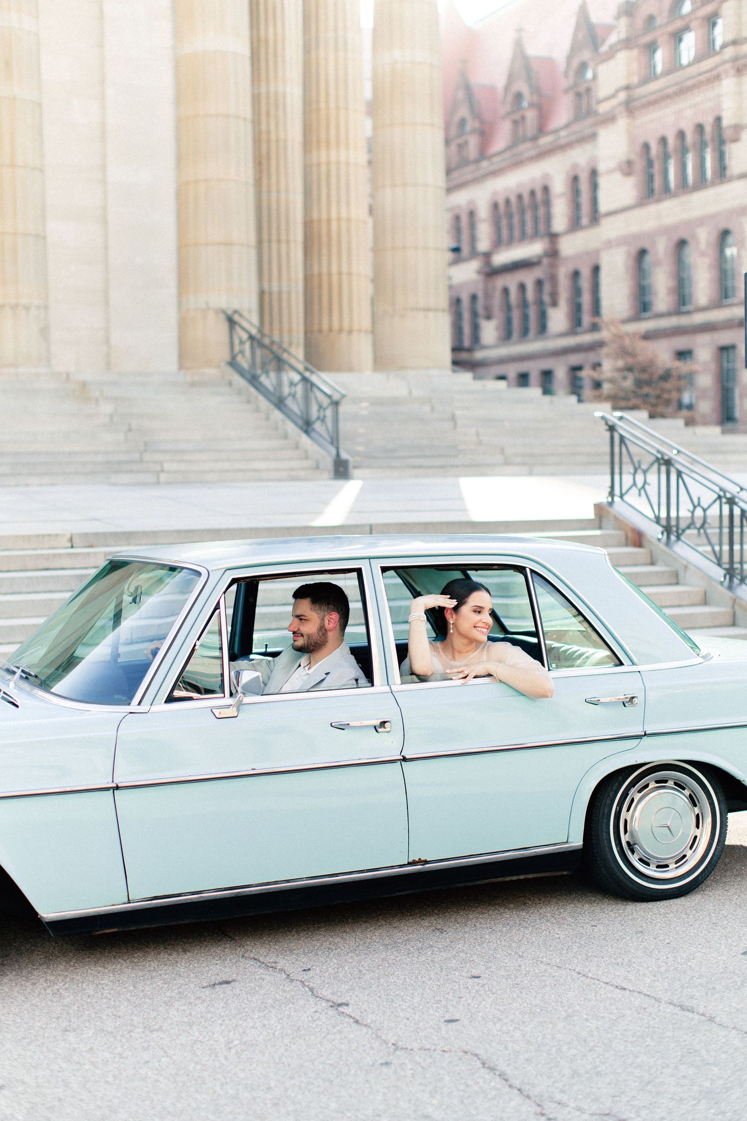 A bride and groom ride in a vintage mint green classic car outside a historic building on their wedding day.