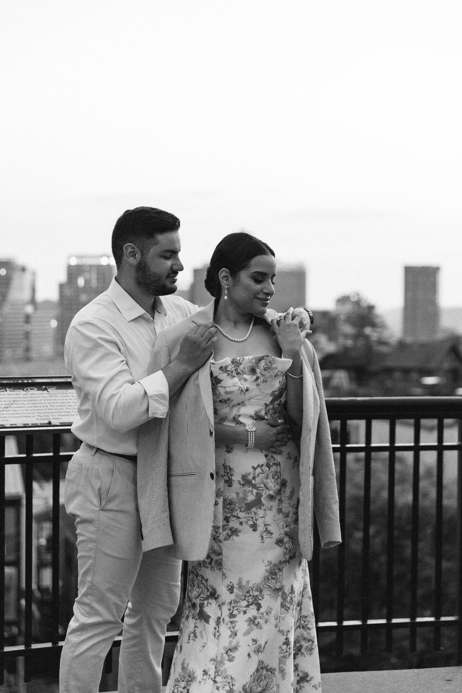 Black and white photo of a couple on a rooftop, man placing jacket on woman's shoulders with city skyline behind them.