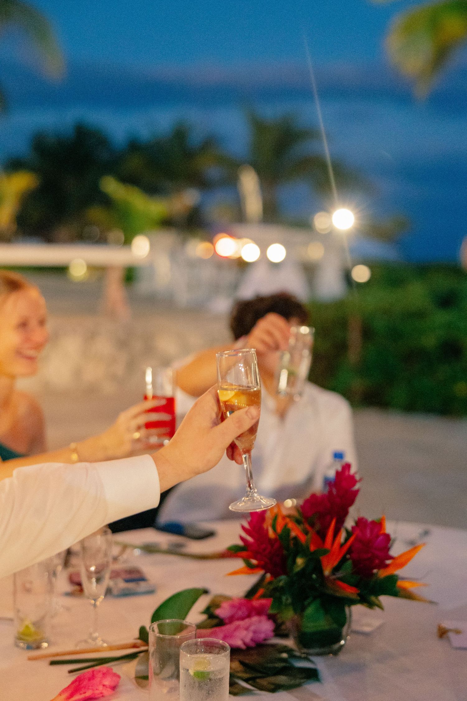 Guests raising champagne glasses in a toast at an elegant outdoor beachside evening wedding reception with tropical floral centerpieces.