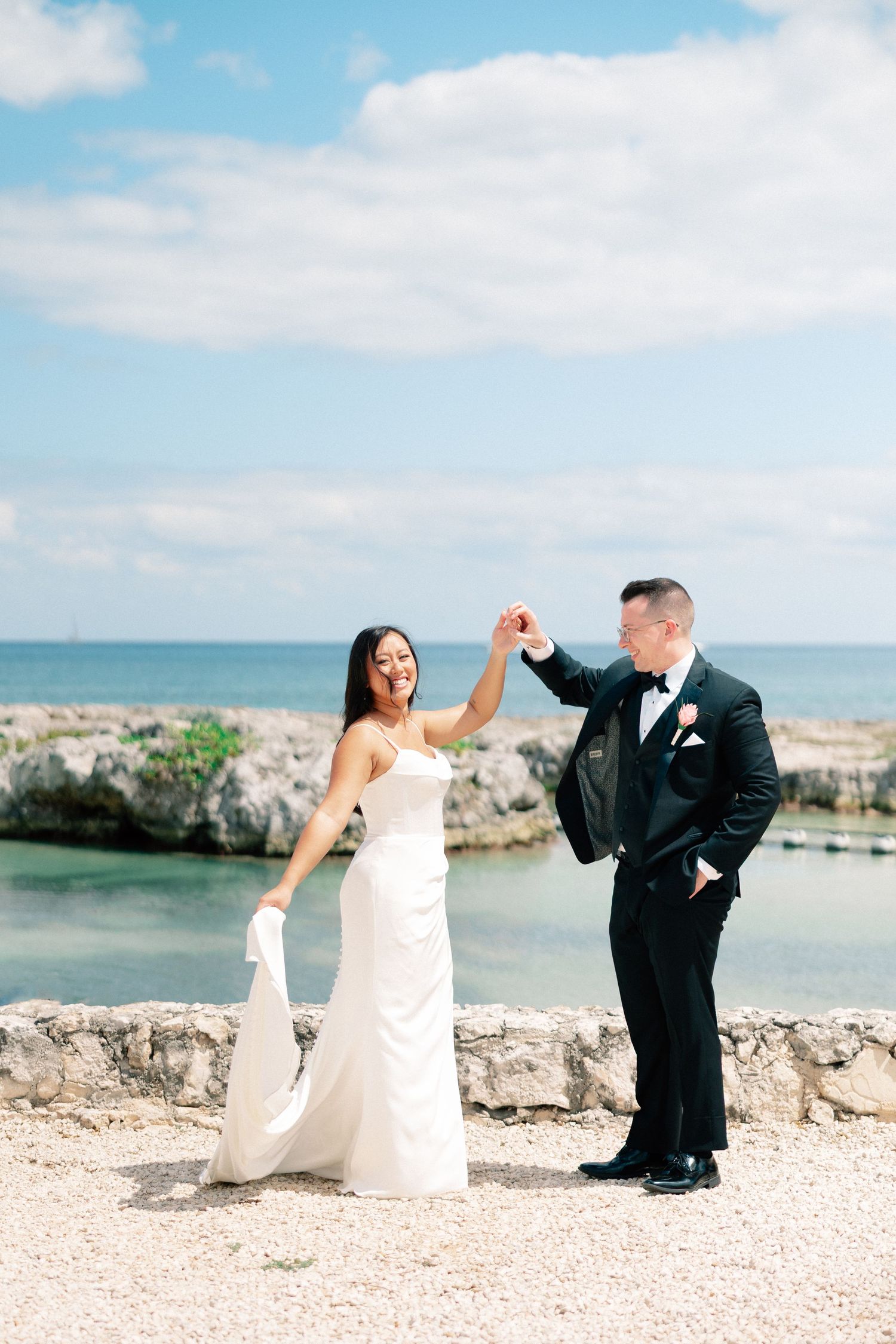 Bride and groom dancing by rocky oceanside, she in white gown, he in black tuxedo, under blue sky.