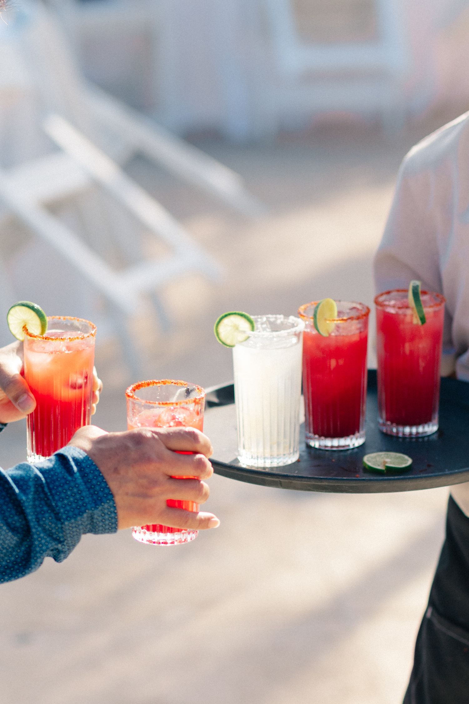 Server holding a tray of red cocktails and a white drink garnished with lime slices at an outdoor event.