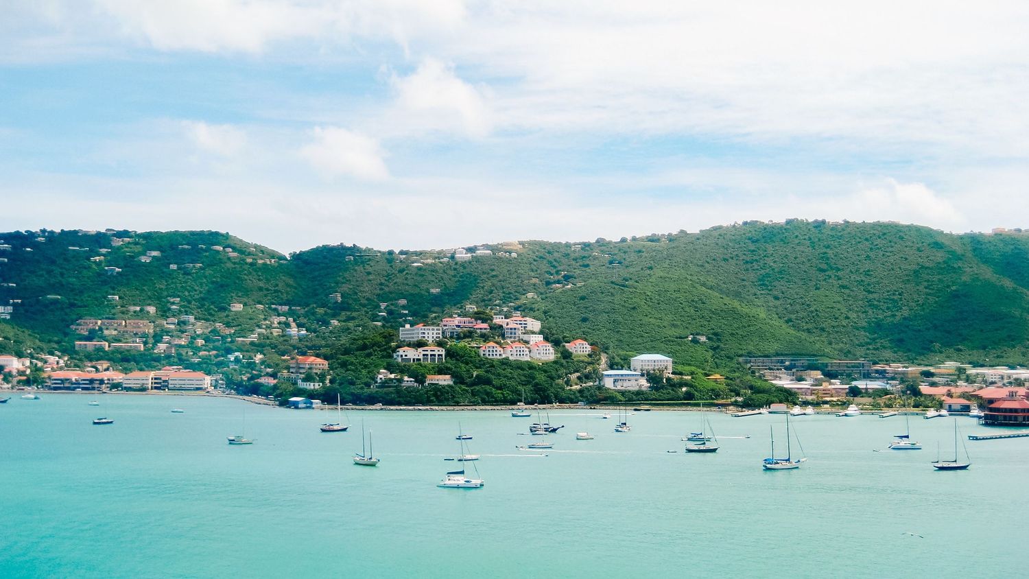 Turquoise Caribbean bay with sailboats anchored near a lush green hillside town under a partly cloudy sky.