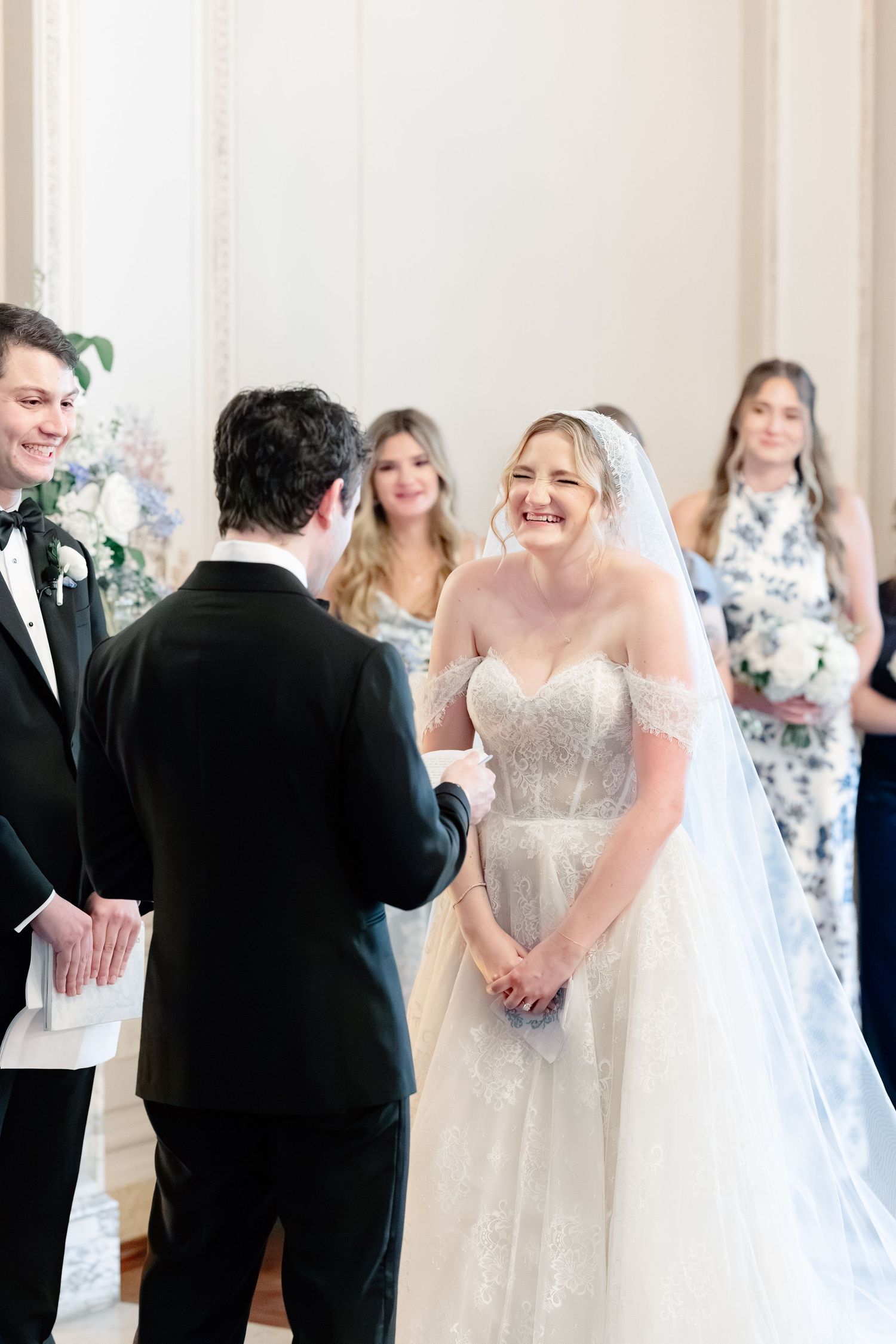 Bride smiling joyfully at groom during wedding ceremony in elegant white venue with bridesmaids in background.