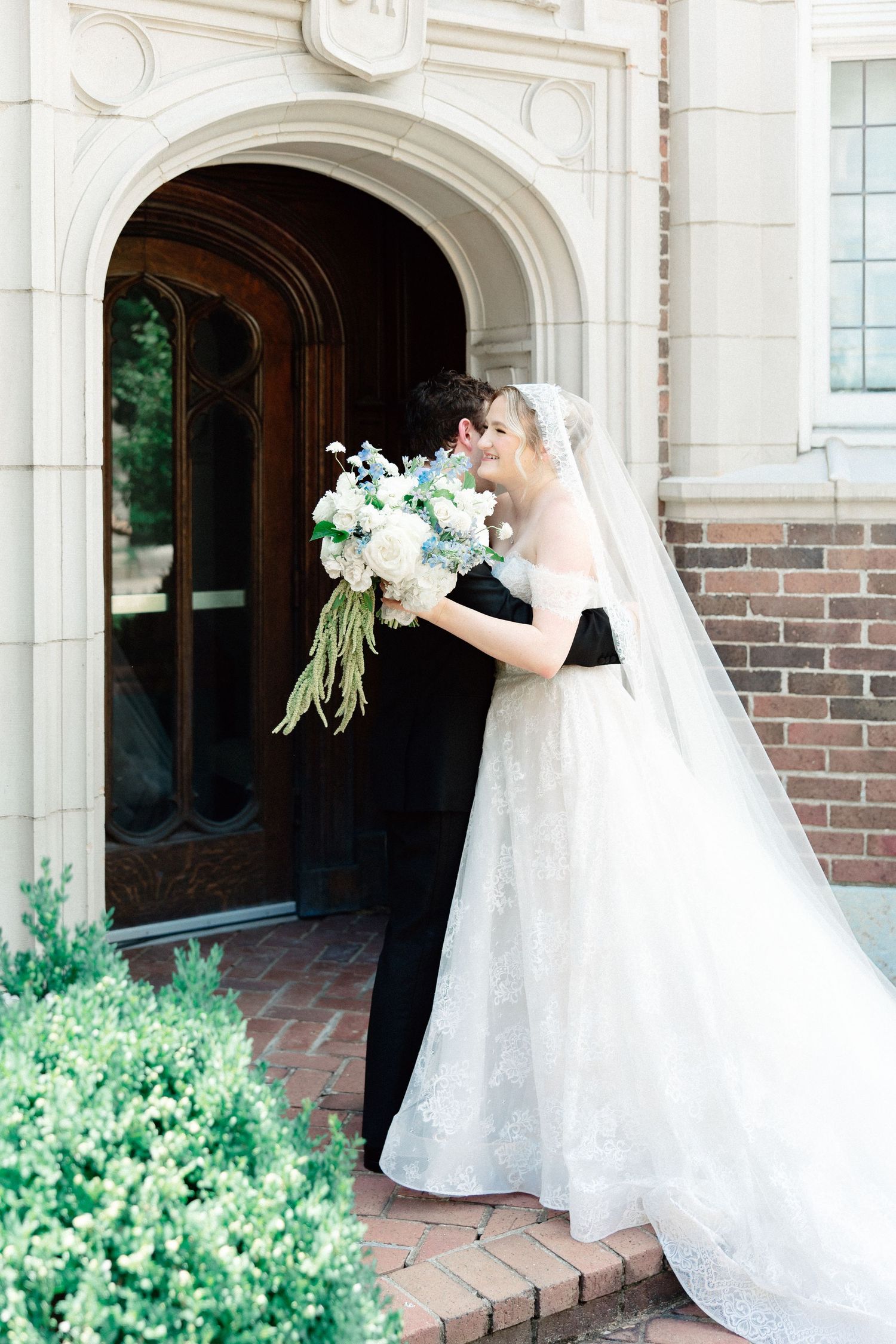 Bride in lace gown and veil kisses groom in black tuxedo holding white bouquet at ornate church entrance.